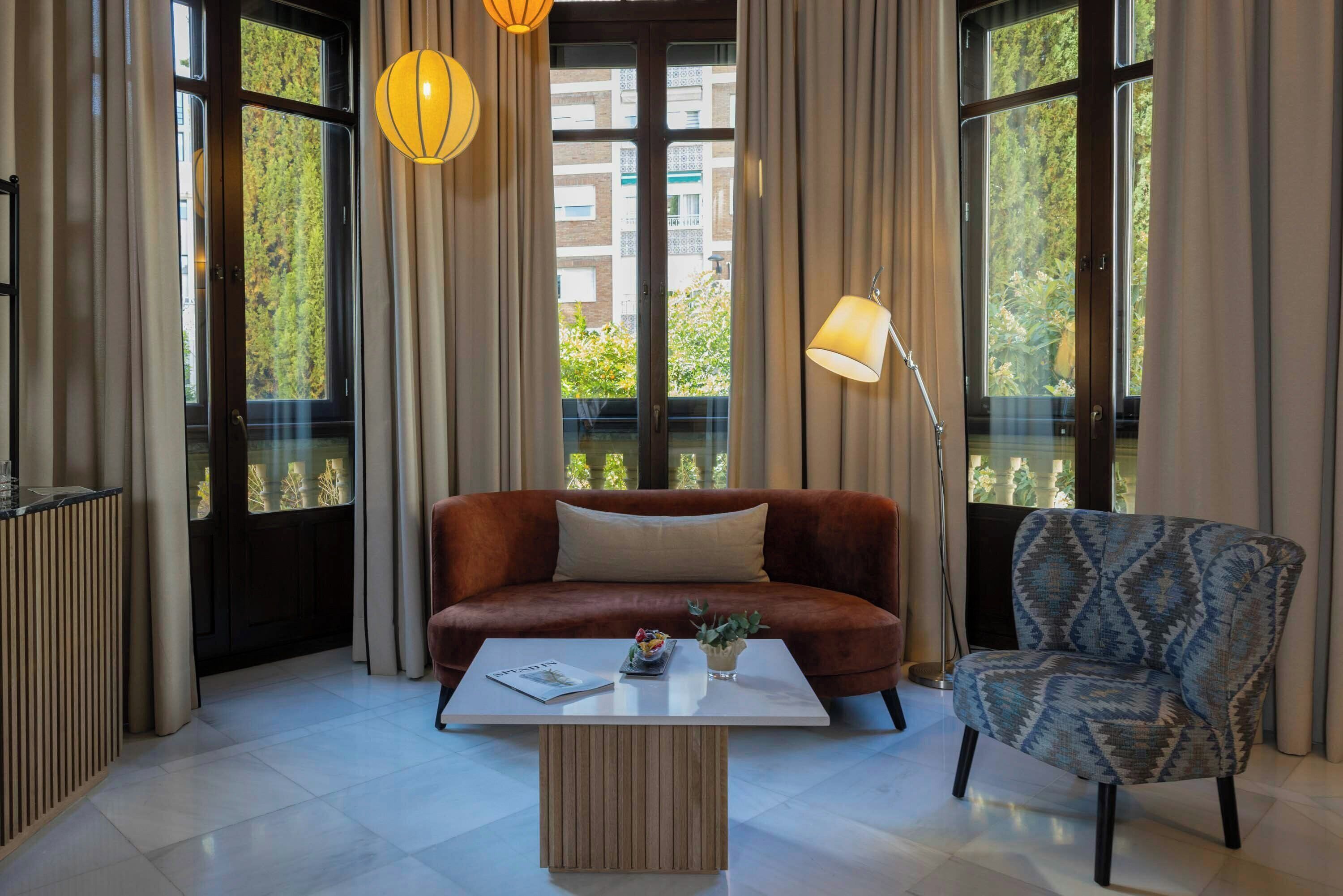 A modern living room with a brown sofa, a patterned armchair, a white coffee table, and large windows with curtains.