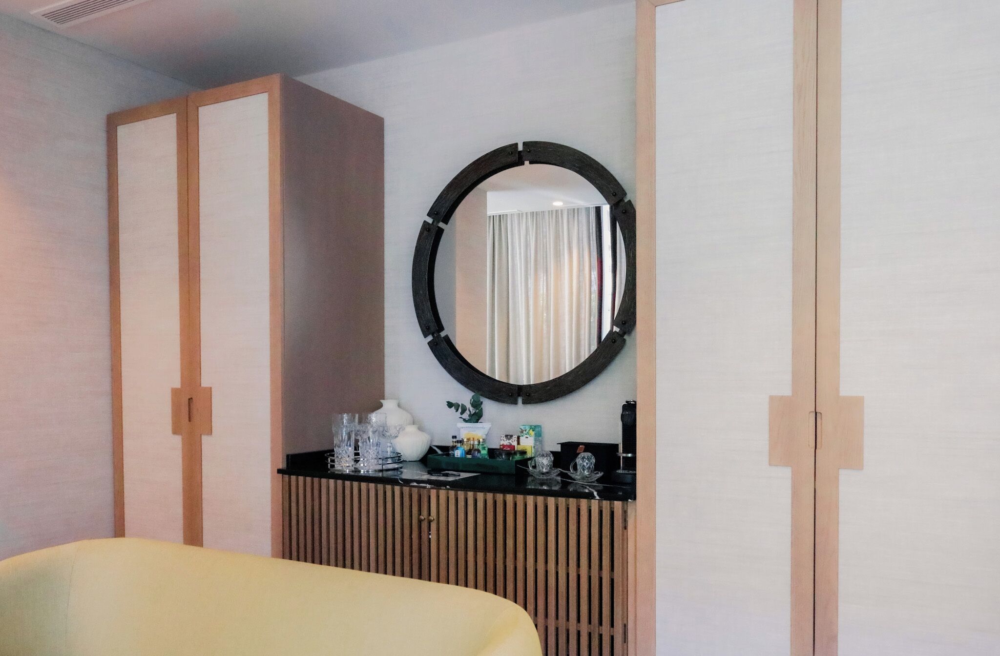 A hotel room with a wooden wardrobe, a round mirror, a small table with toiletries, and a yellow armchair.