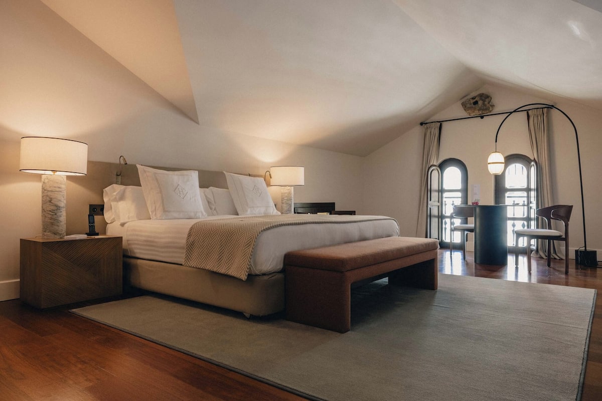 Ein Schlafzimmer mit Bett, Nachttischen, einer Bank und Blick auf einen Essbereich.