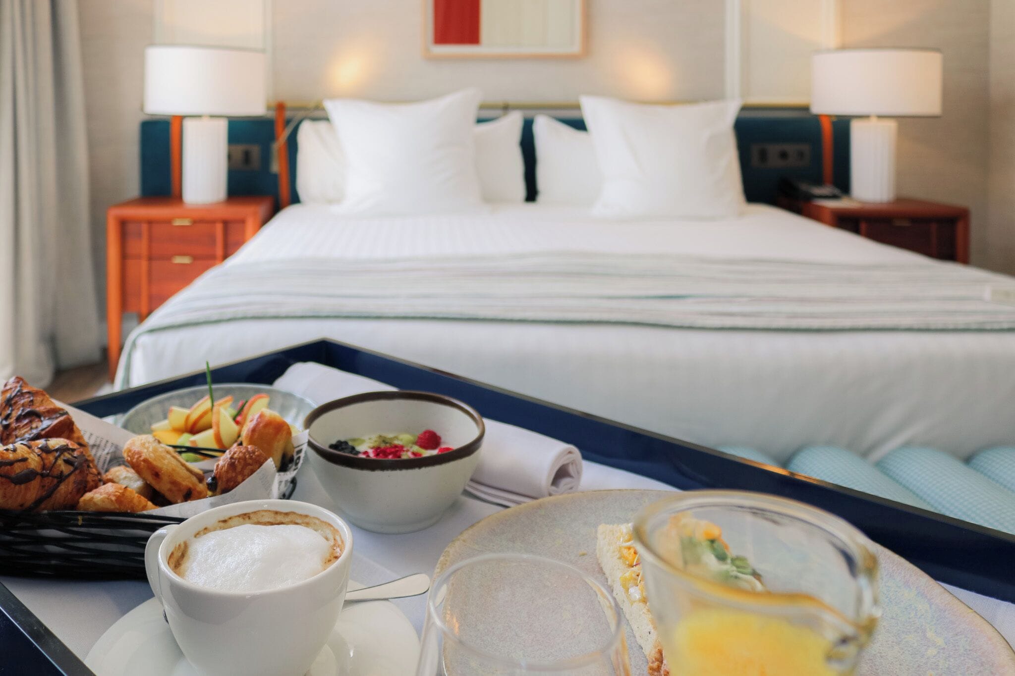 A hotel room with a neatly made bed, a breakfast tray with croissants, fruit, and coffee, and two bedside lamps.
