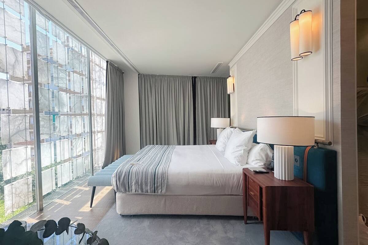 Ein Hotelzimmer mit einem großen Bett, zwei Nachttischen, einem Fenster mit Vorhängen und Blick nach außen.