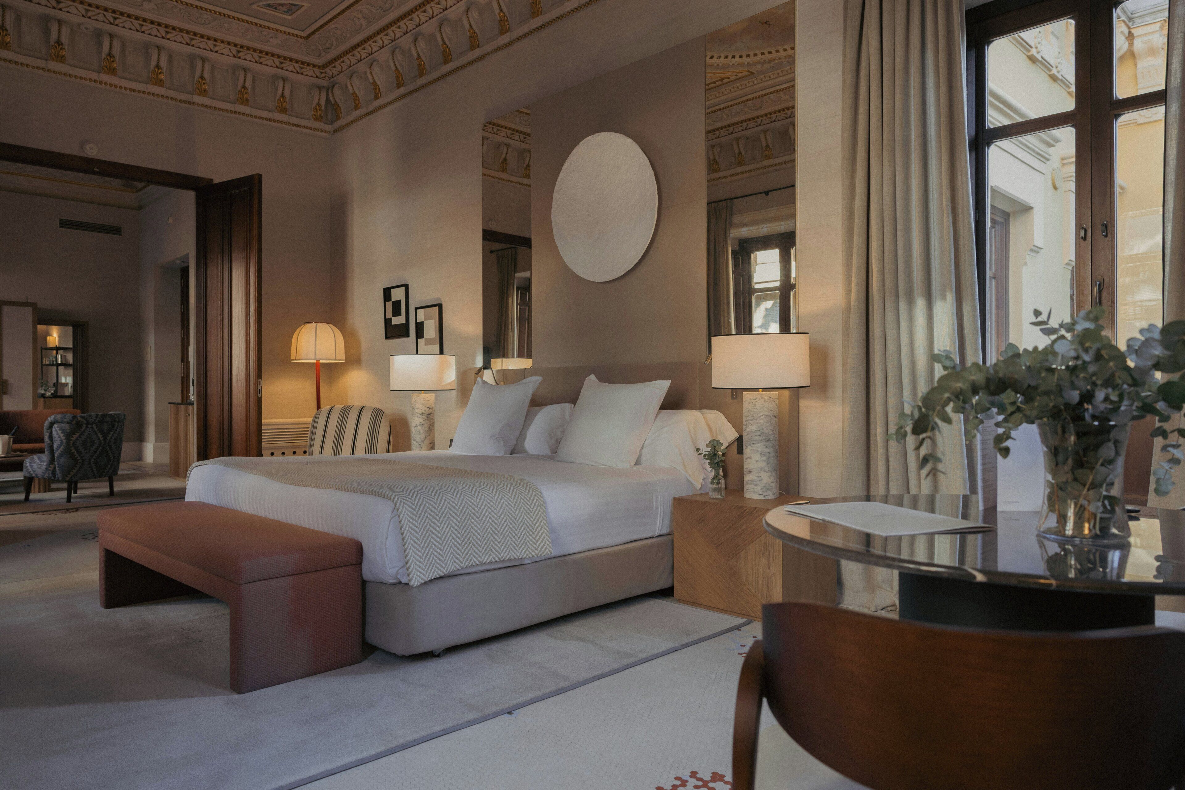 Une chambre d’hôtel avec un grand lit, des lampes de chevet, un miroir rond et une petite table sur laquelle est posé un vase.