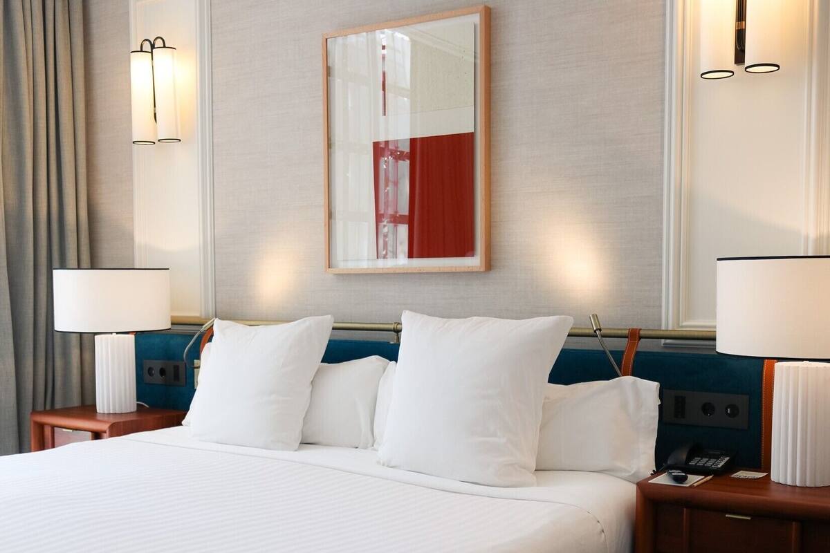 Ein Hotelzimmer mit einem großen Bett, zwei Nachttischen inklusive Lampen, einem Spiegel an der Wand und einem Fenster mit Vorhängen.