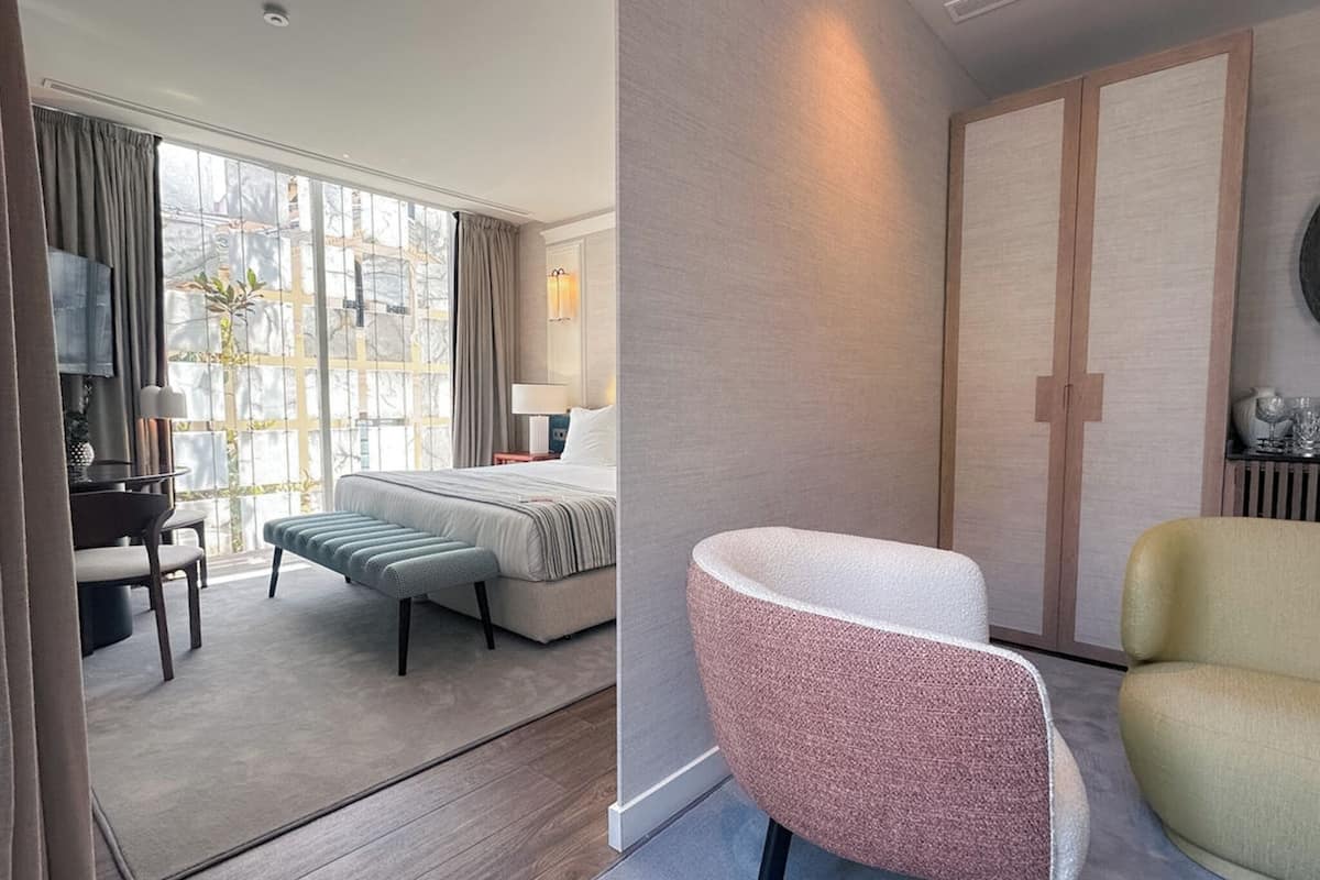 Ein modernes Hotelzimmer mit einem Bett, einer Sitzecke mit einer Bank und Sesseln, einem Schreibtisch und einem großen Fenster mit Vorhängen.