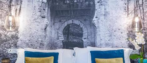 A room with a bed, two blue and gold pillows, a nightstand with a candle, and a wall mural of a stone archway.