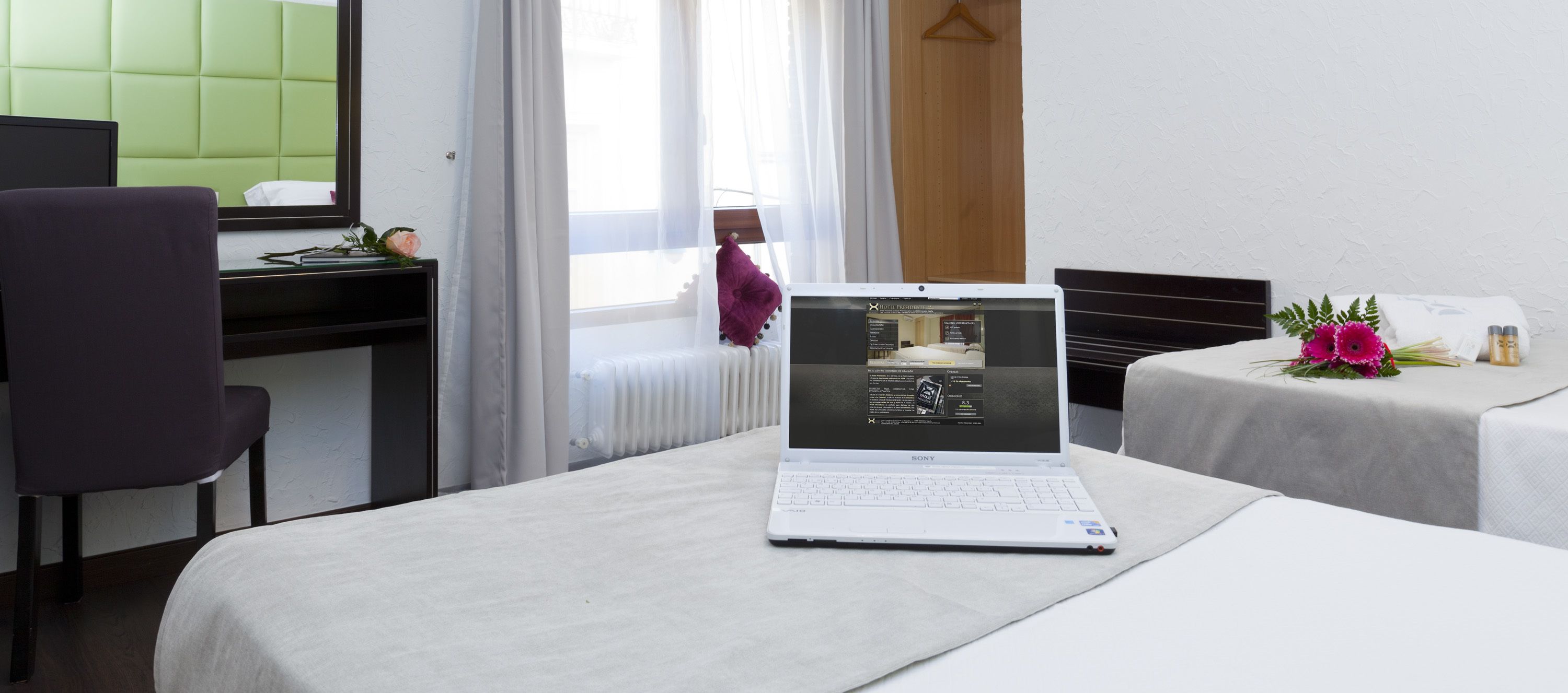 A hotel room with a bed, a desk with a laptop, a chair, and a small table with a bouquet of flowers.