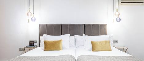 Una cama doble con cabecera gris, ropa de cama blanca y dos almohadas doradas. Hay mesitas de noche con una lámpara pequeña y un libro.