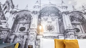 Une chambre à coucher ornée d’une grande fresque représentant un bâtiment historique, un lit avec des coussins blancs et moutarde, un bureau avec une chaise, et une petite table avec une bougie.