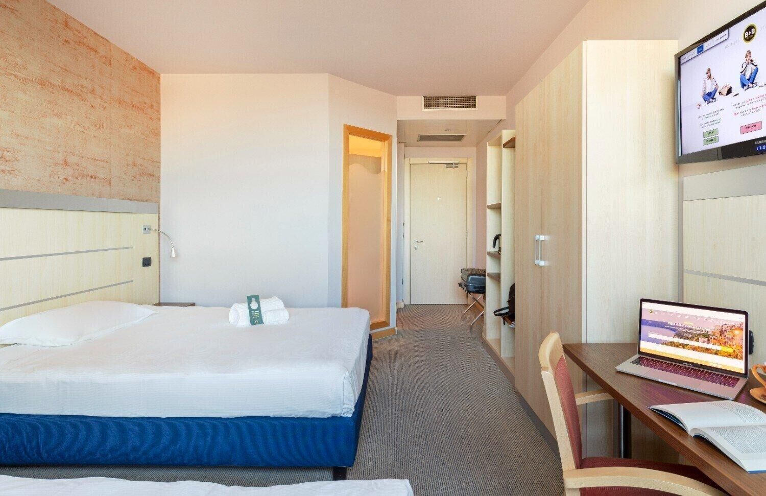 Ein Hotelzimmer mit einem Bett, einem Schreibtisch mit einem Laptop und einem Fernseher, der ein Programm zeigt.
