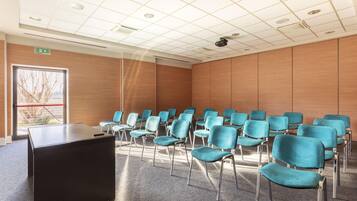 Un salón de conferencias con filas de sillas color turquesa, un atril negro y un ventanal con vistas a árboles.