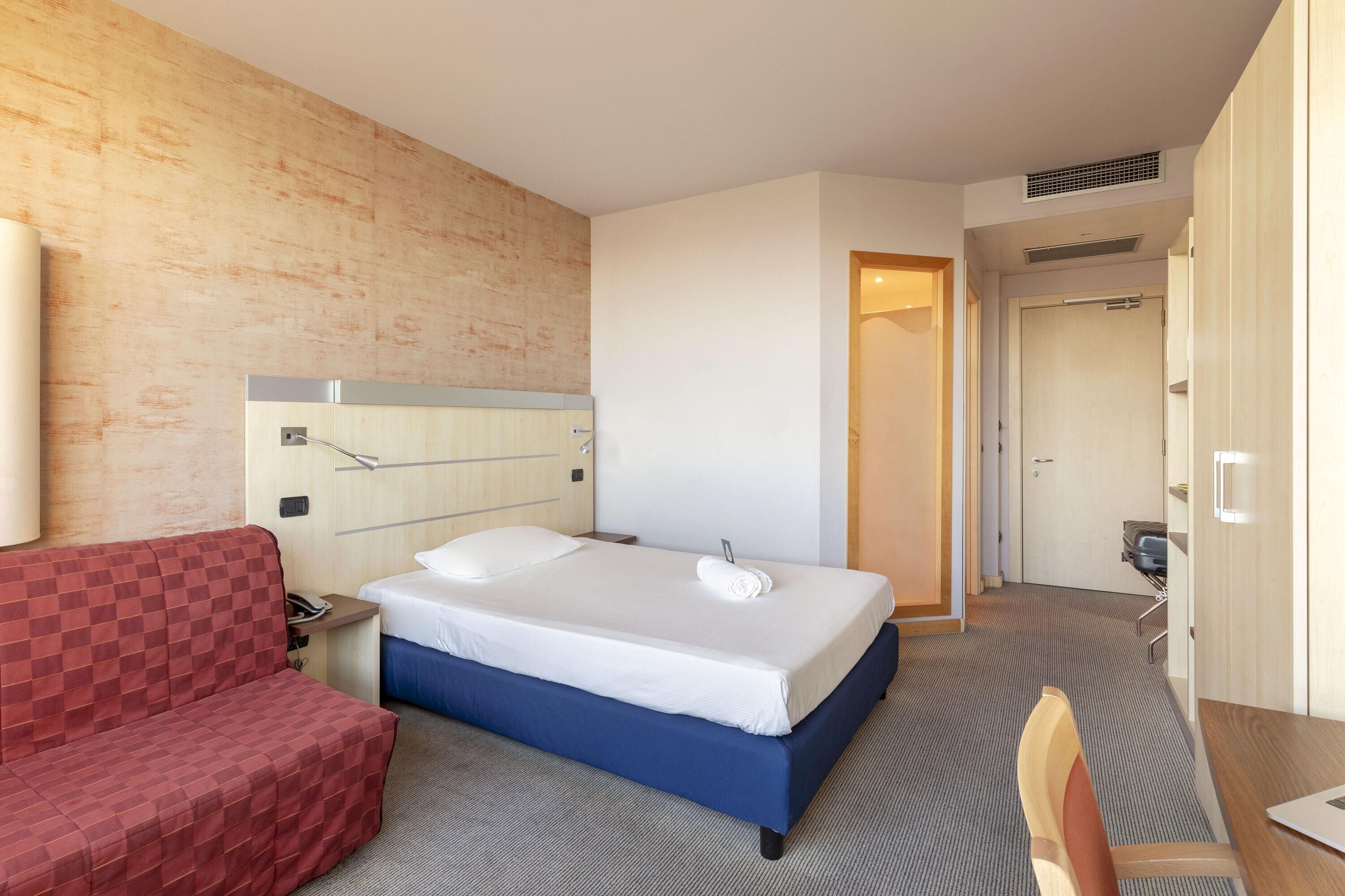 Une chambre d’hôtel avec un lit, un canapé rouge à carreaux, un bureau et un miroir.
