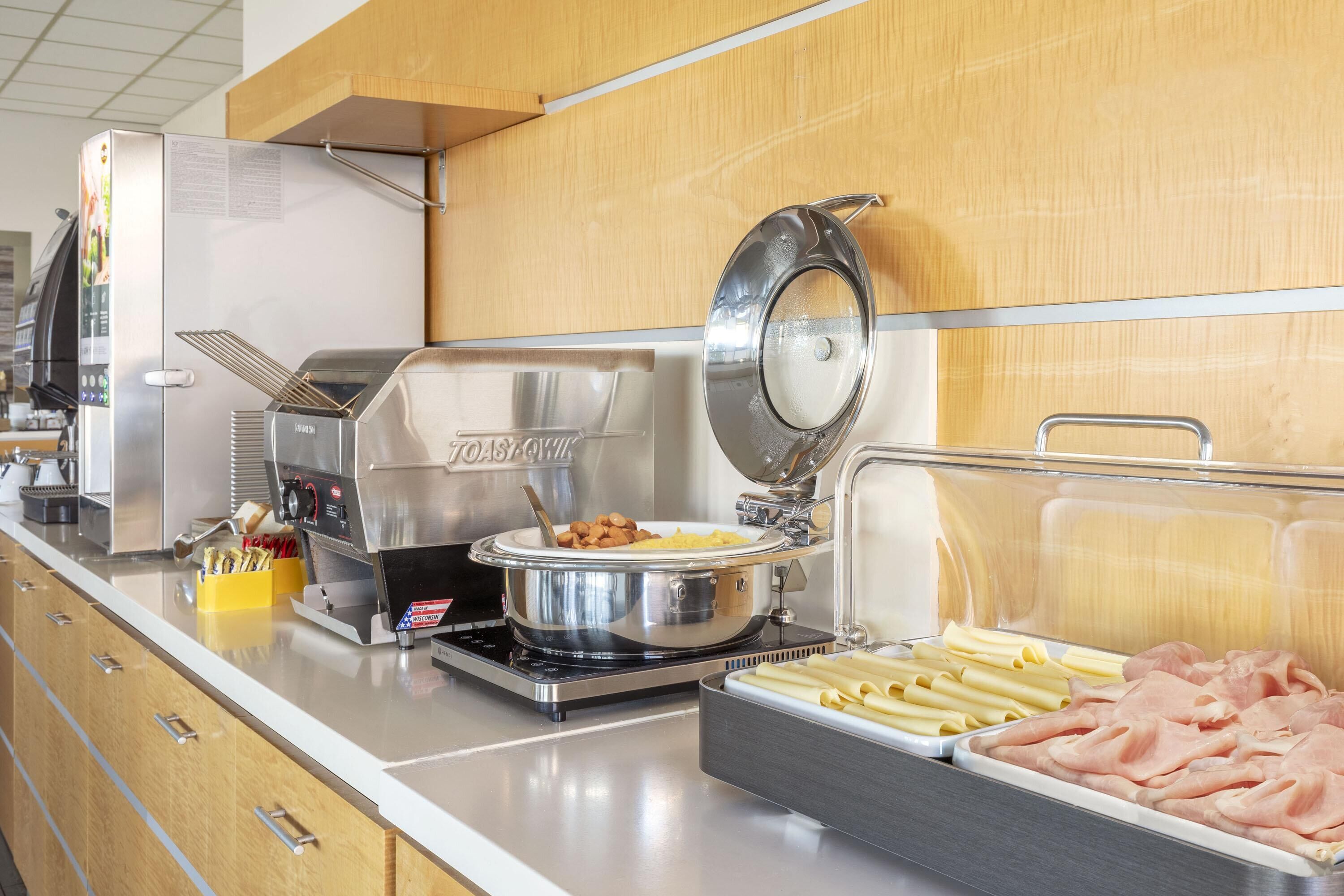 A buffet setup with a coffee machine, a hot plate, and various food items including pasta and sliced meats.
