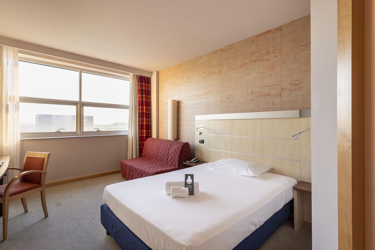 Een hotelkamer met een groot bed, een bureau met stoel, een rode bank en een raam met gordijnen.