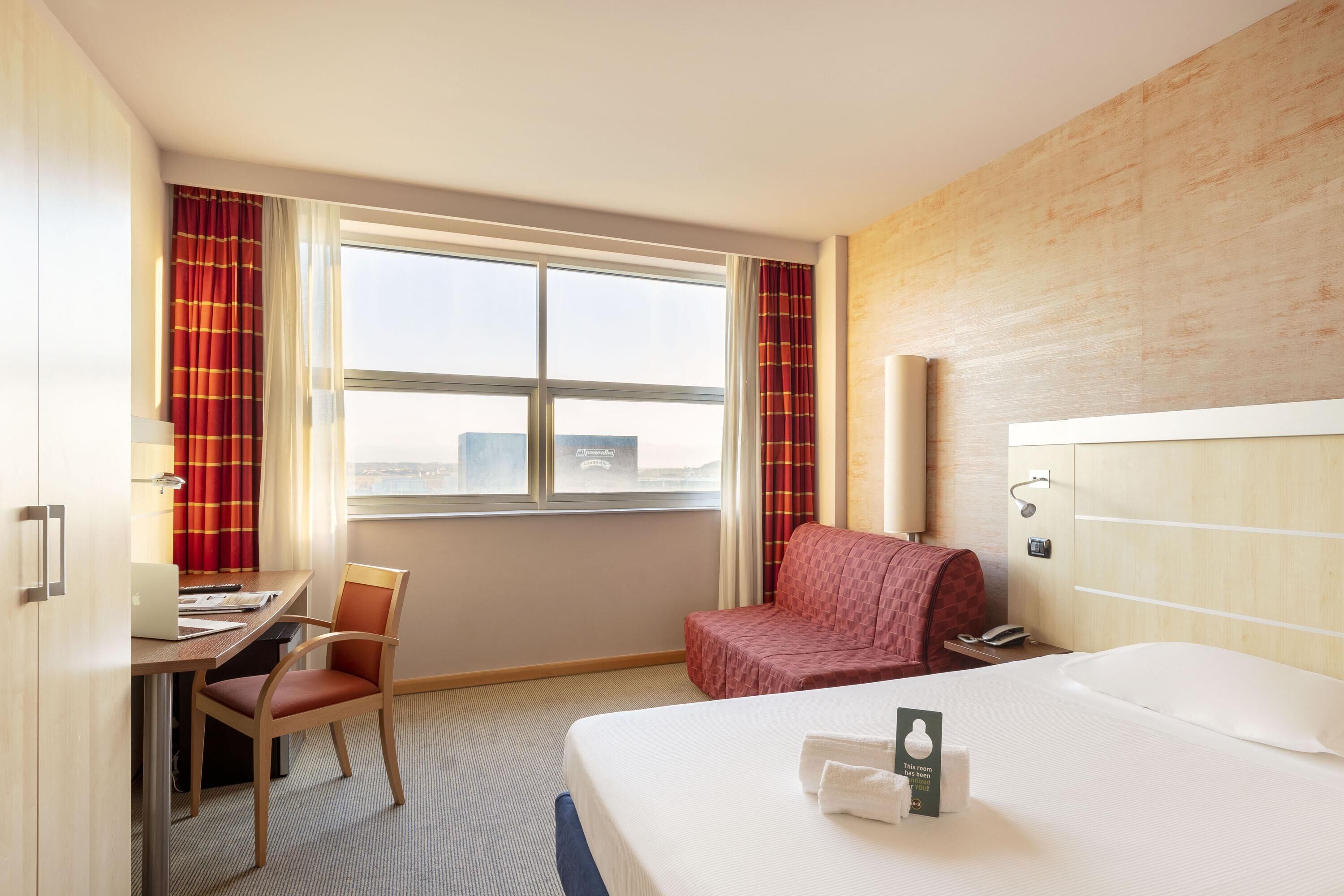 Habitación de hotel con cama, escritorio con computadora portátil, silla, sofá rojo y ventana con cortinas.