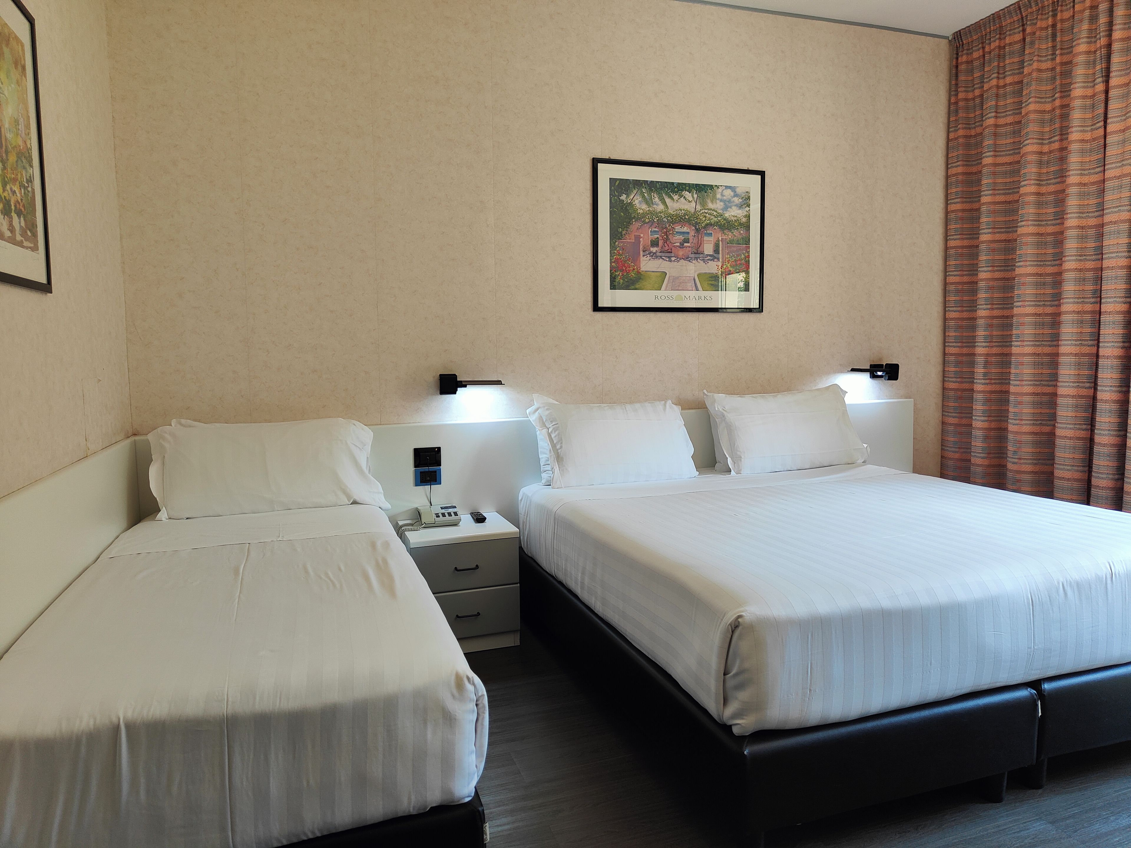 A hotel room with two beds, a nightstand, a wall-mounted lamp, and a framed picture.