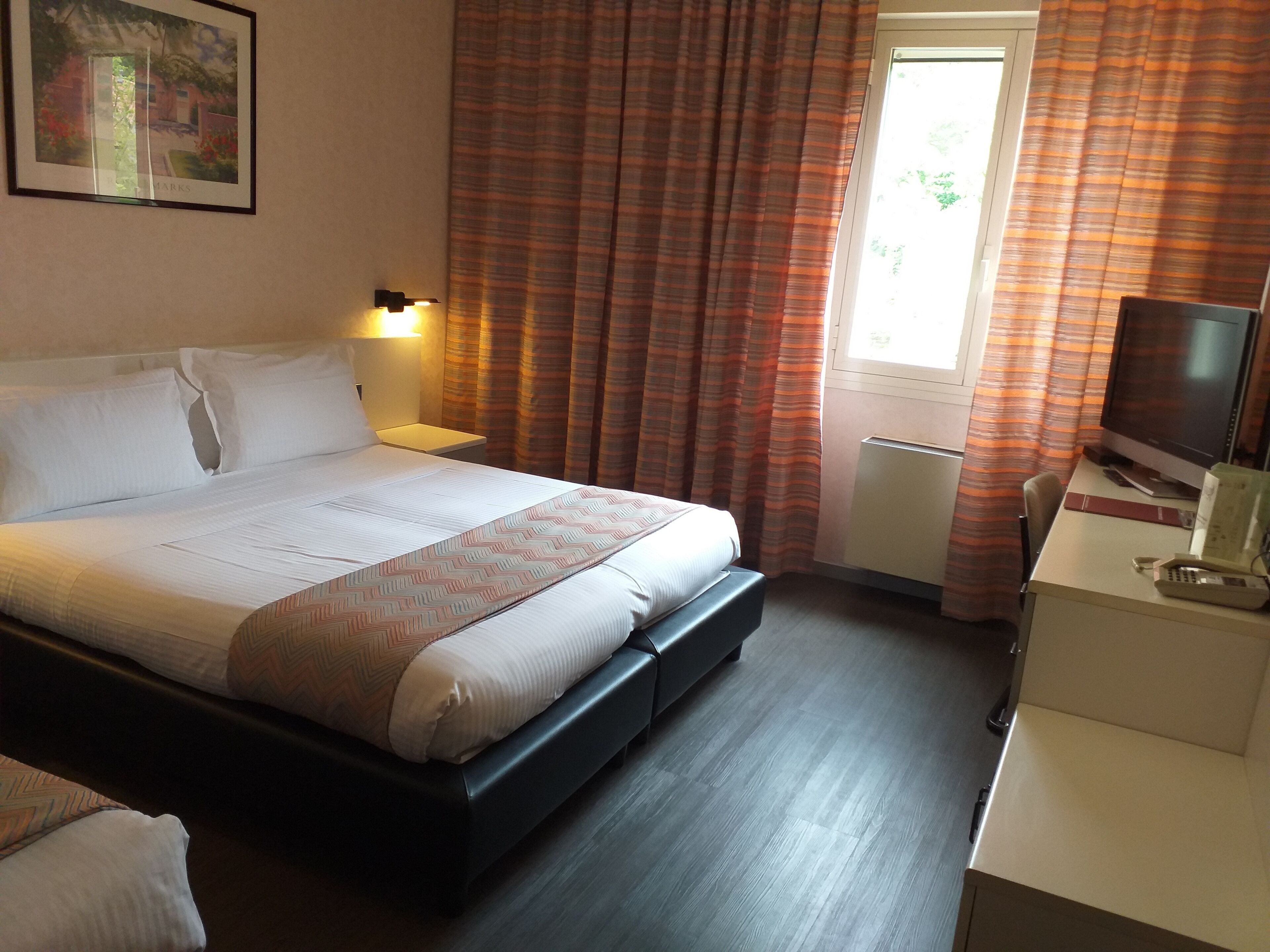 A hotel room with a bed, a desk with a computer, a window with curtains, and a wall-mounted picture.