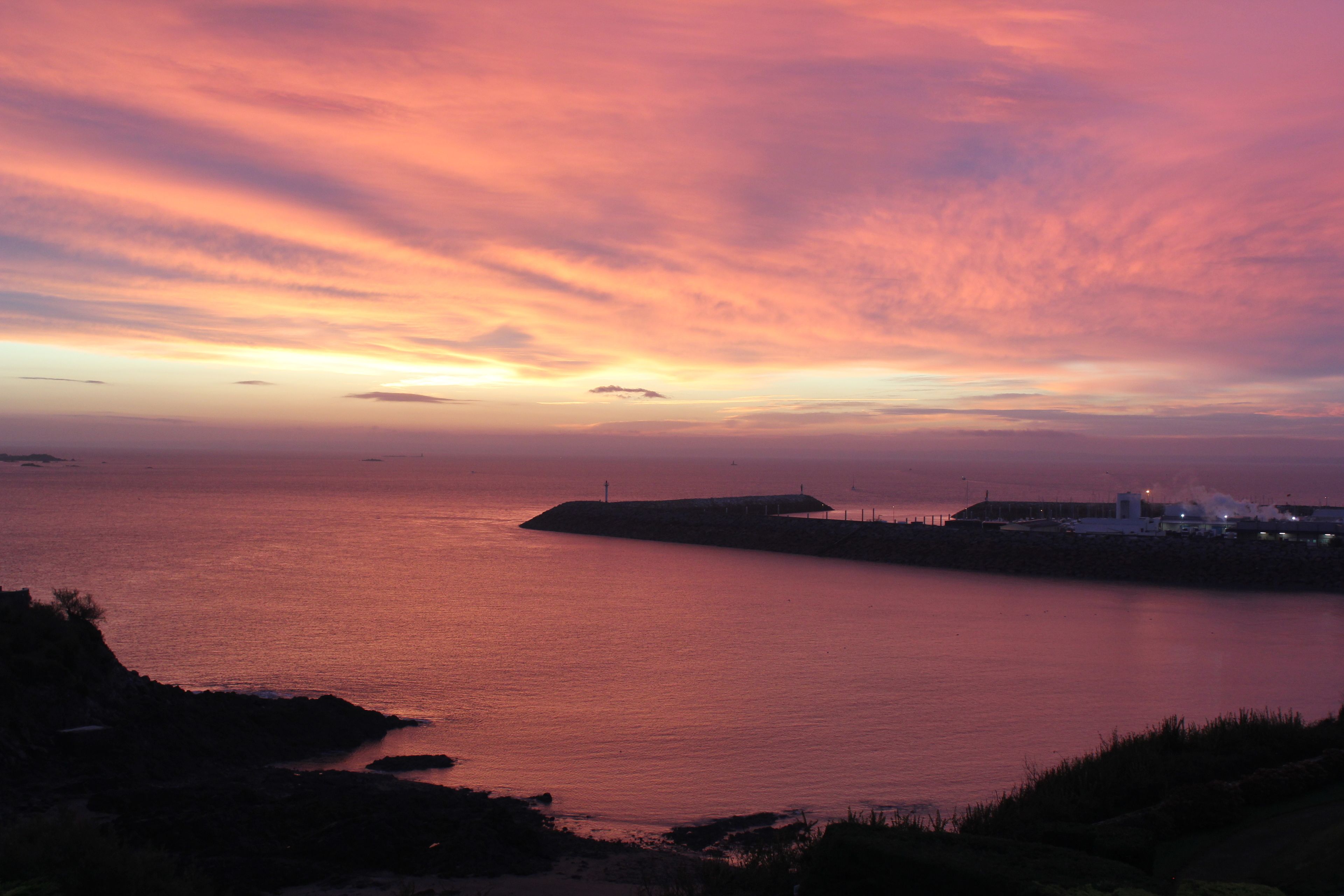 Une vue sur le coucher de soleil en bord de mer, avec une jetée et des bâtiments éloignés.