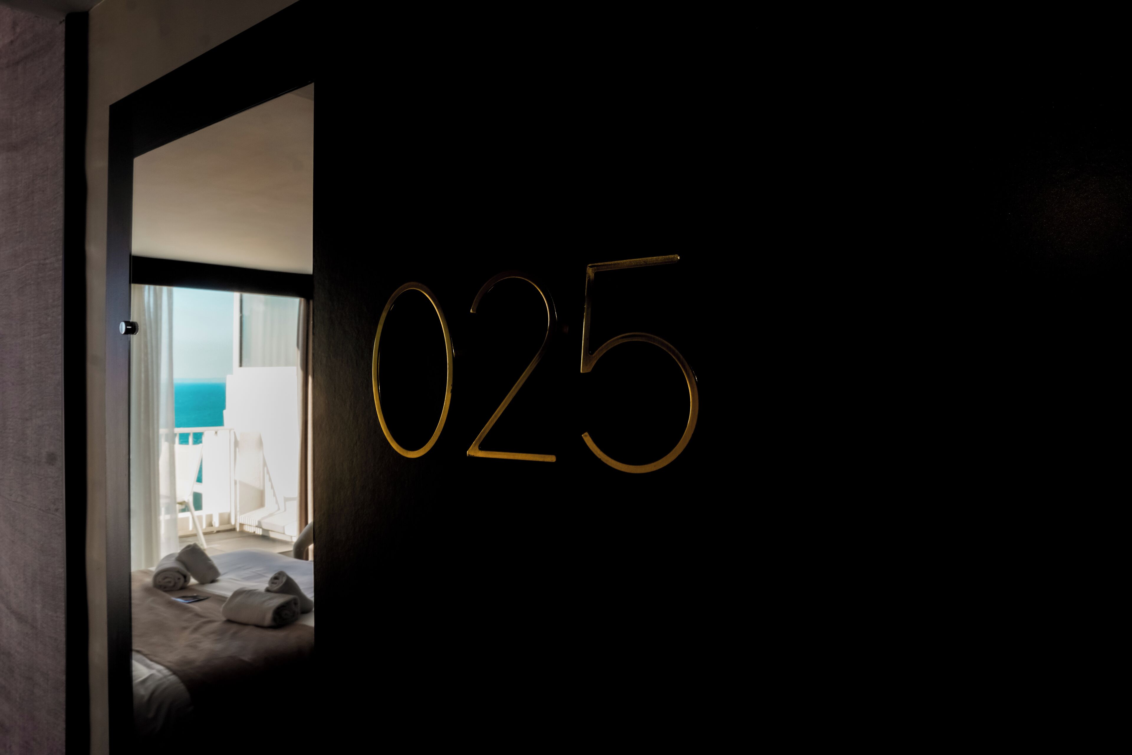 Une pièce sombre, avec le numéro 025 peint sur le mur, un lit recouvert de serviettes enroulées et une vue sur un balcon avec un ciel bleu.