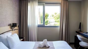 A hotel room with a bed, a desk, a chair, a window with curtains, and a view of greenery outside.