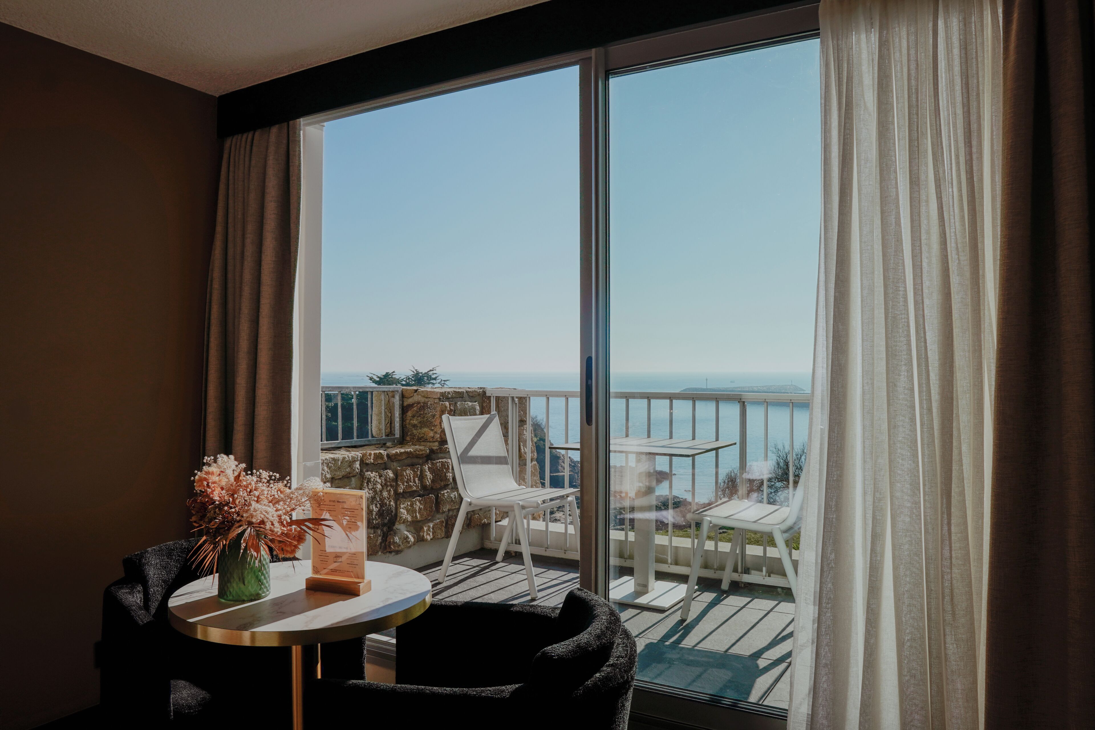 A balcony with a view of the sea, a table with a vase of flowers, and a chair.
