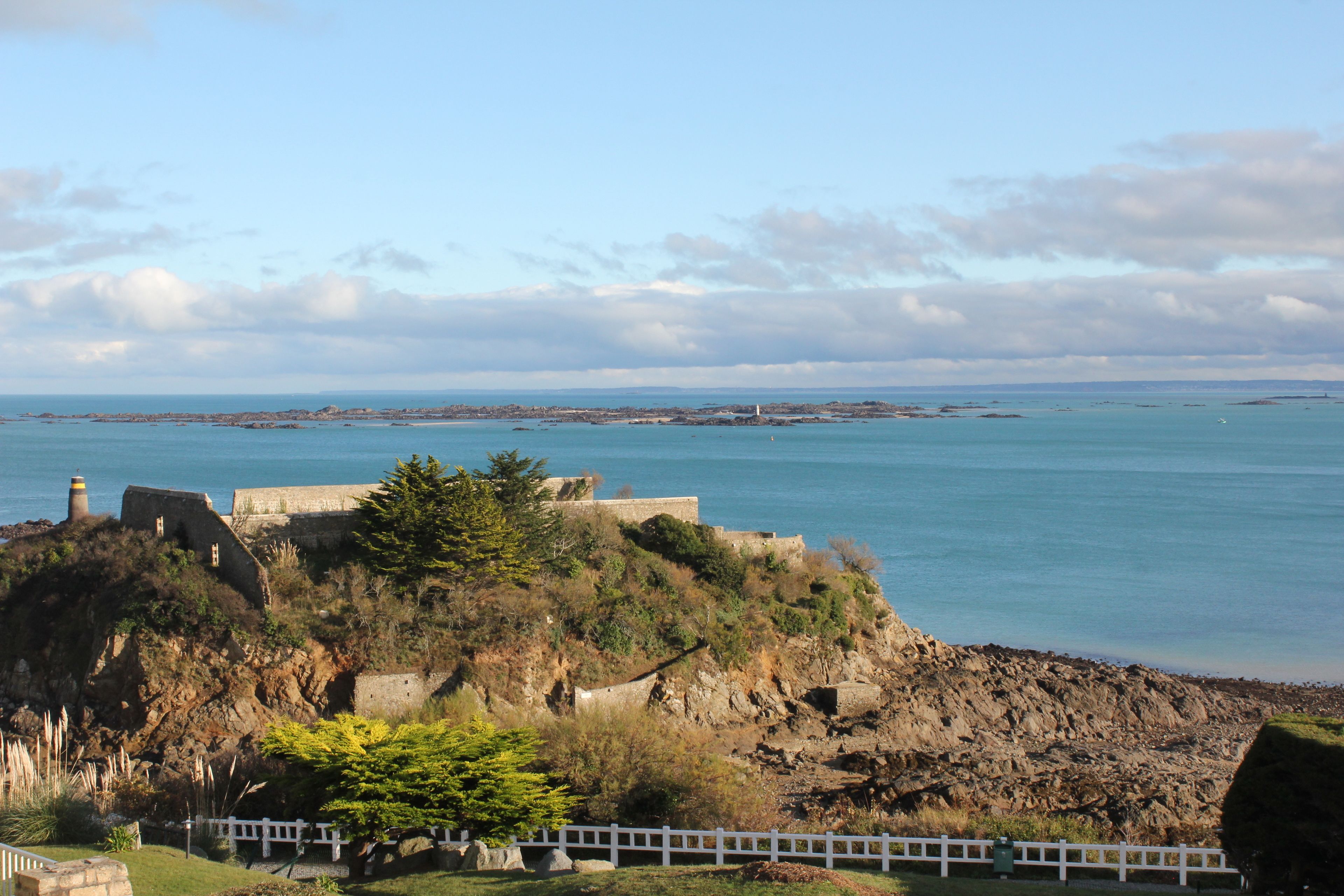 A coastal fortification with a lighthouse, rocky terrain, and a white fence.