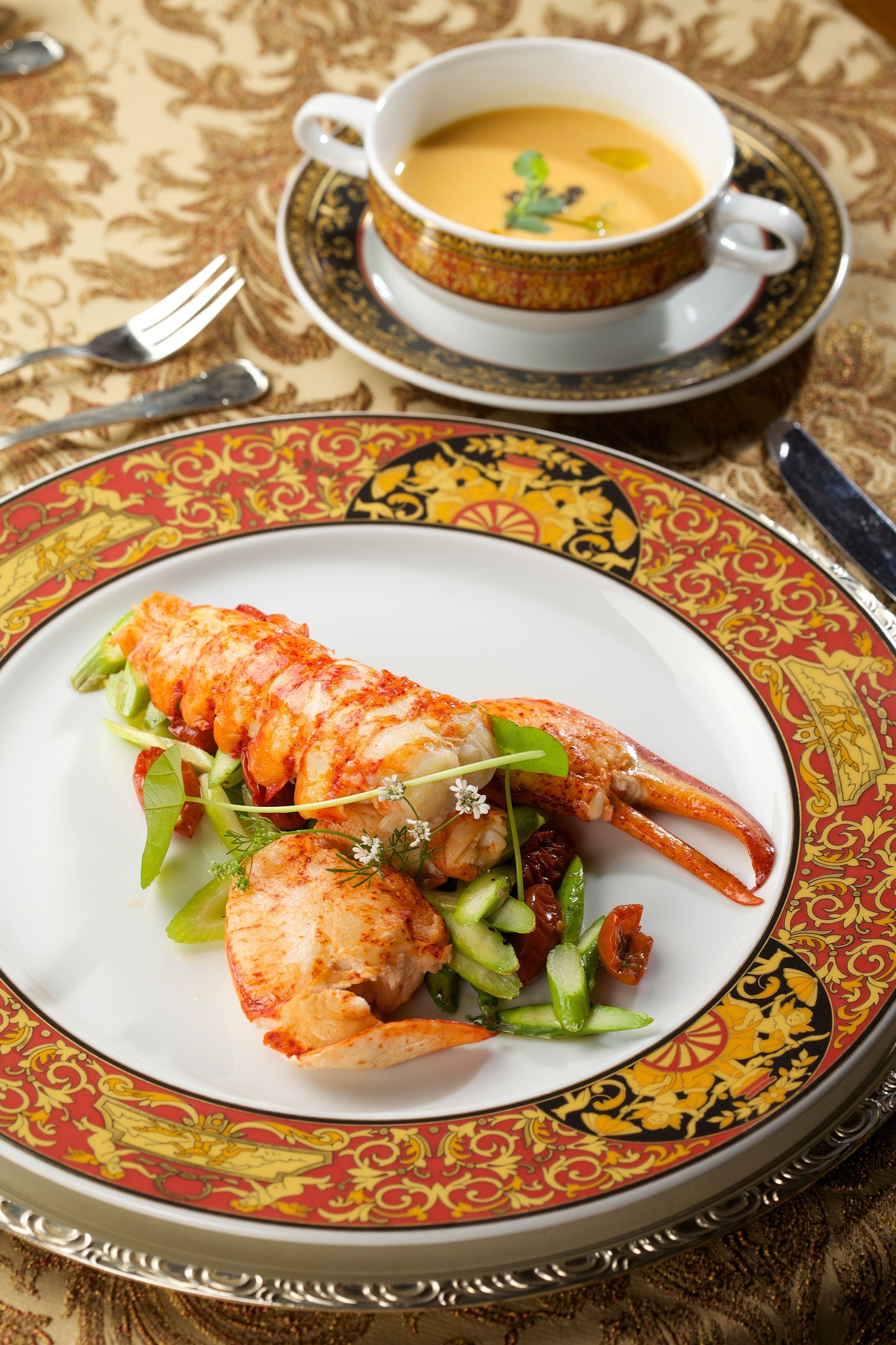 Un plato de langosta sobre un plato decorativo con un mantel estampado y una taza de sopa al fondo.