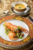 Un plato de langosta sobre un plato decorativo con un mantel estampado y una taza de sopa al fondo.