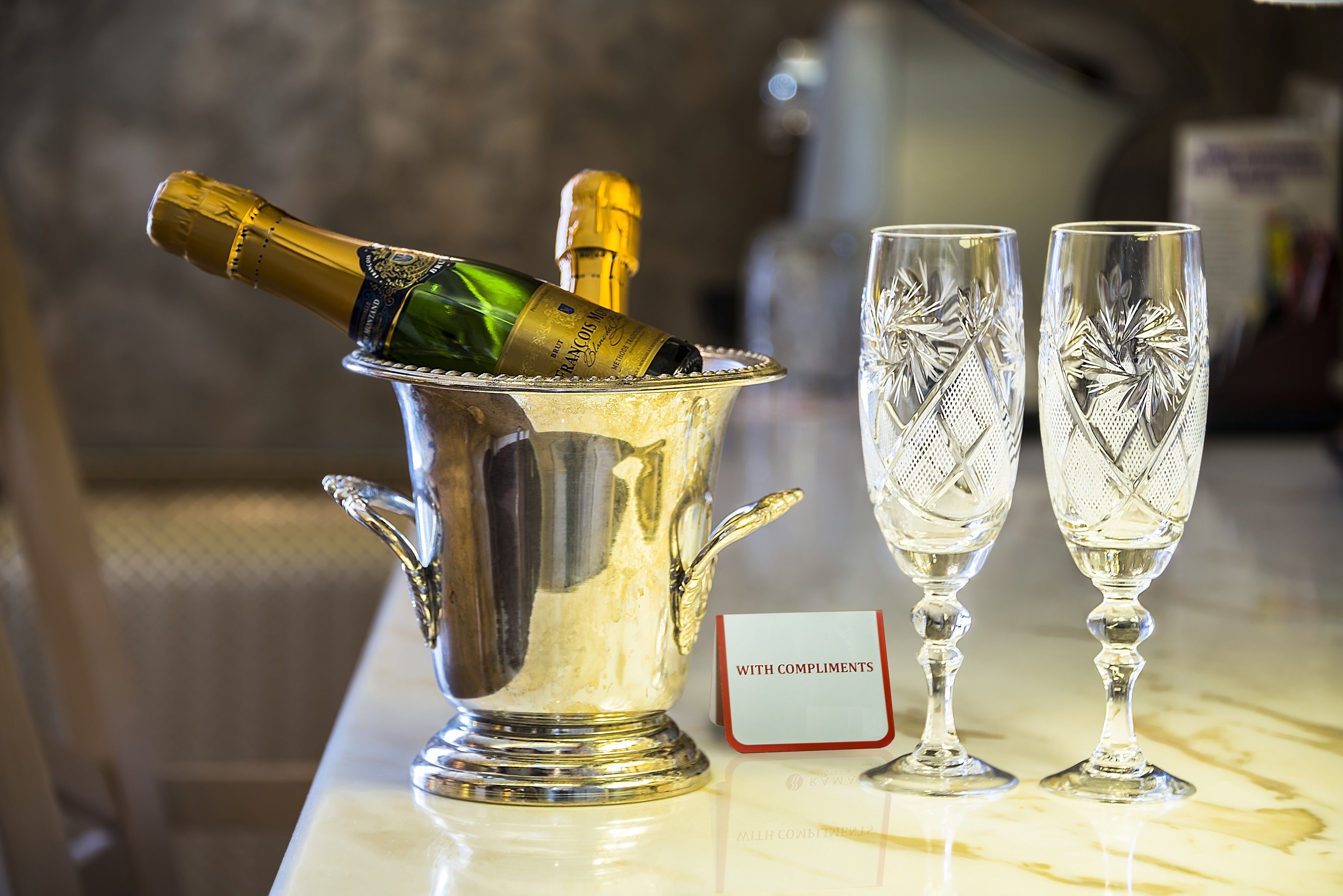 A champagne bottle in a silver ice bucket with two crystal glasses beside it, and a sign that reads "WITH COMPLIMENTS".