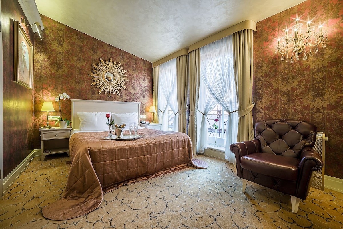 Una habitación de hotel con una cama grande, una zona de estar con un sillón de cuero, un espejo decorativo y una araña.