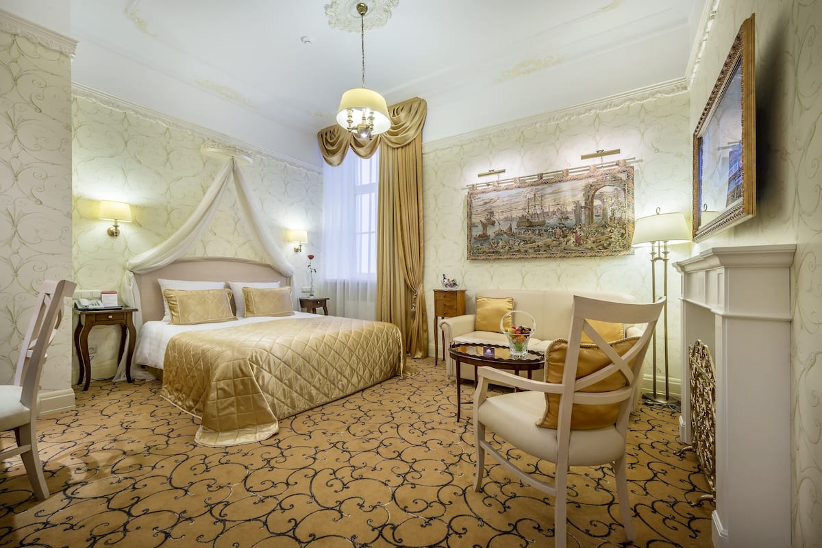 Habitación de hotel con una cama grande, una zona de estar con sofá y sillones, una mesita con un jarrón de flores y un cuadro en la pared.