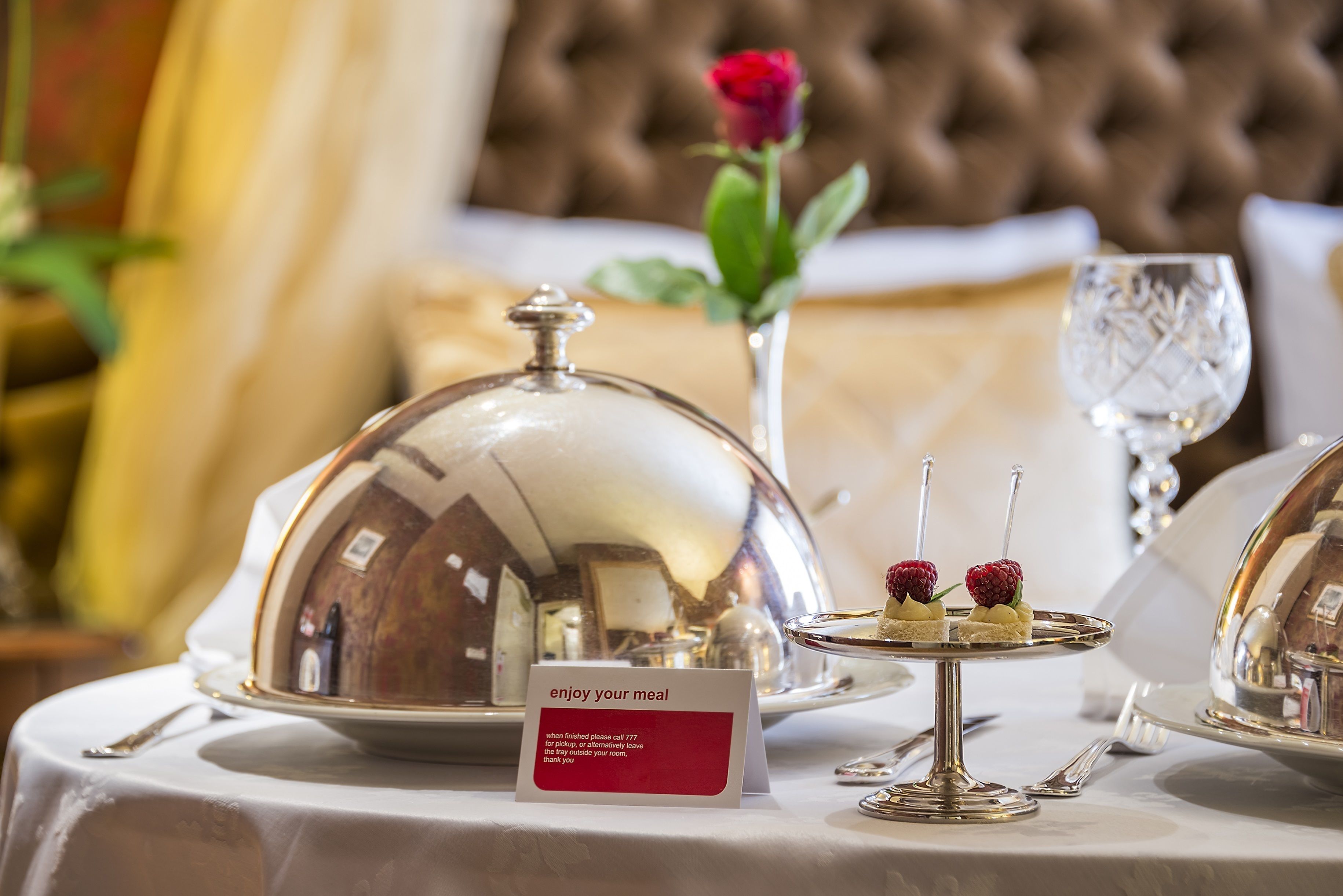 A dining table with a silver dome cover, a small dessert stand with two desserts, a glass of water, and a rose in the background.