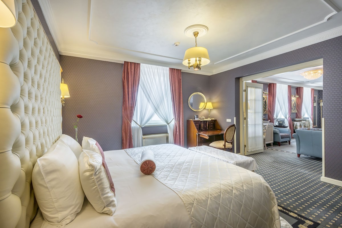 Habitación de hotel con una cama grande, una zona de estar con una silla y una mesa, un espejo y vistas al pasillo.