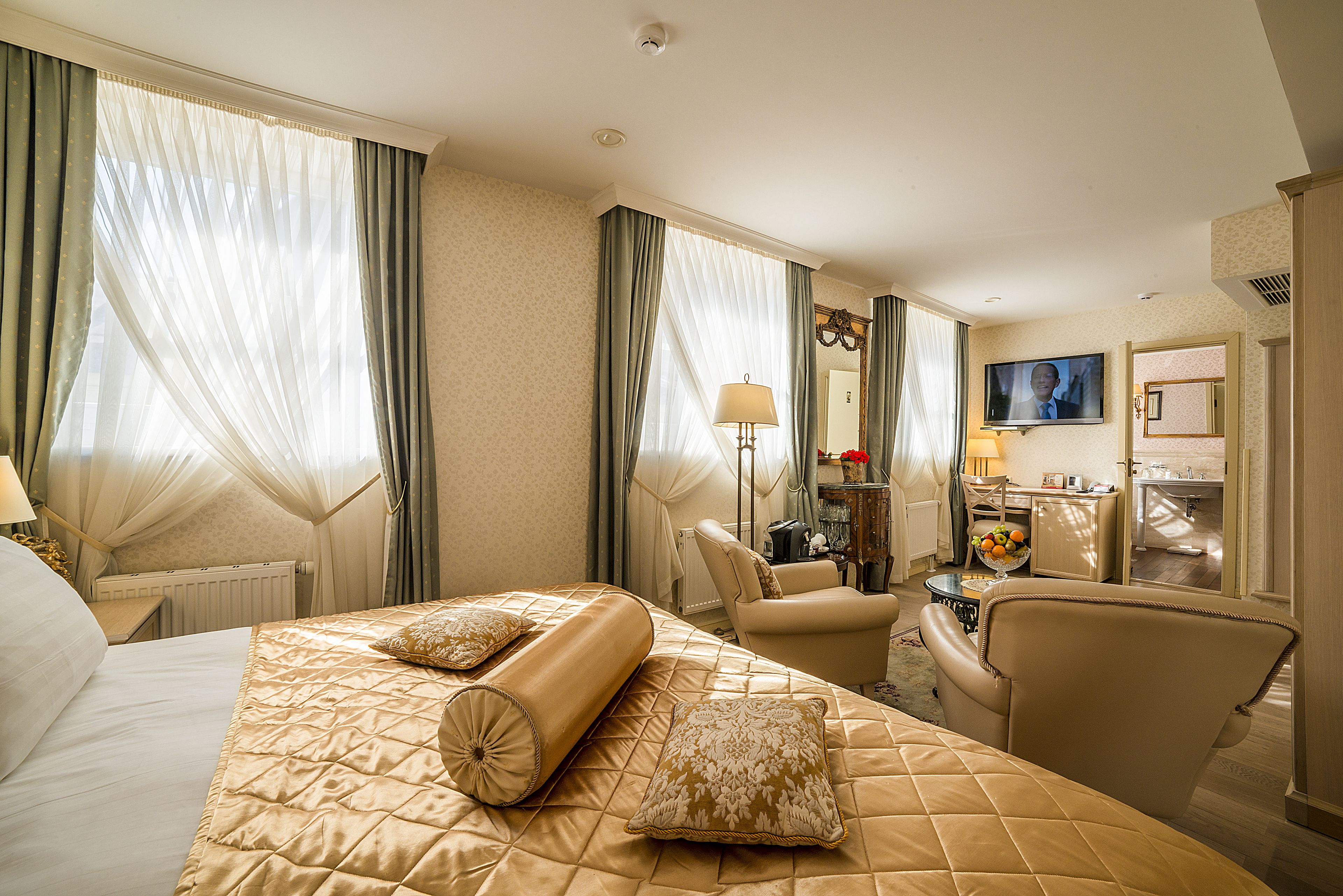 Habitación de hotel con una cama grande, una zona de estar con una silla y un escritorio, televisión y baño visible a través de una puerta abierta.