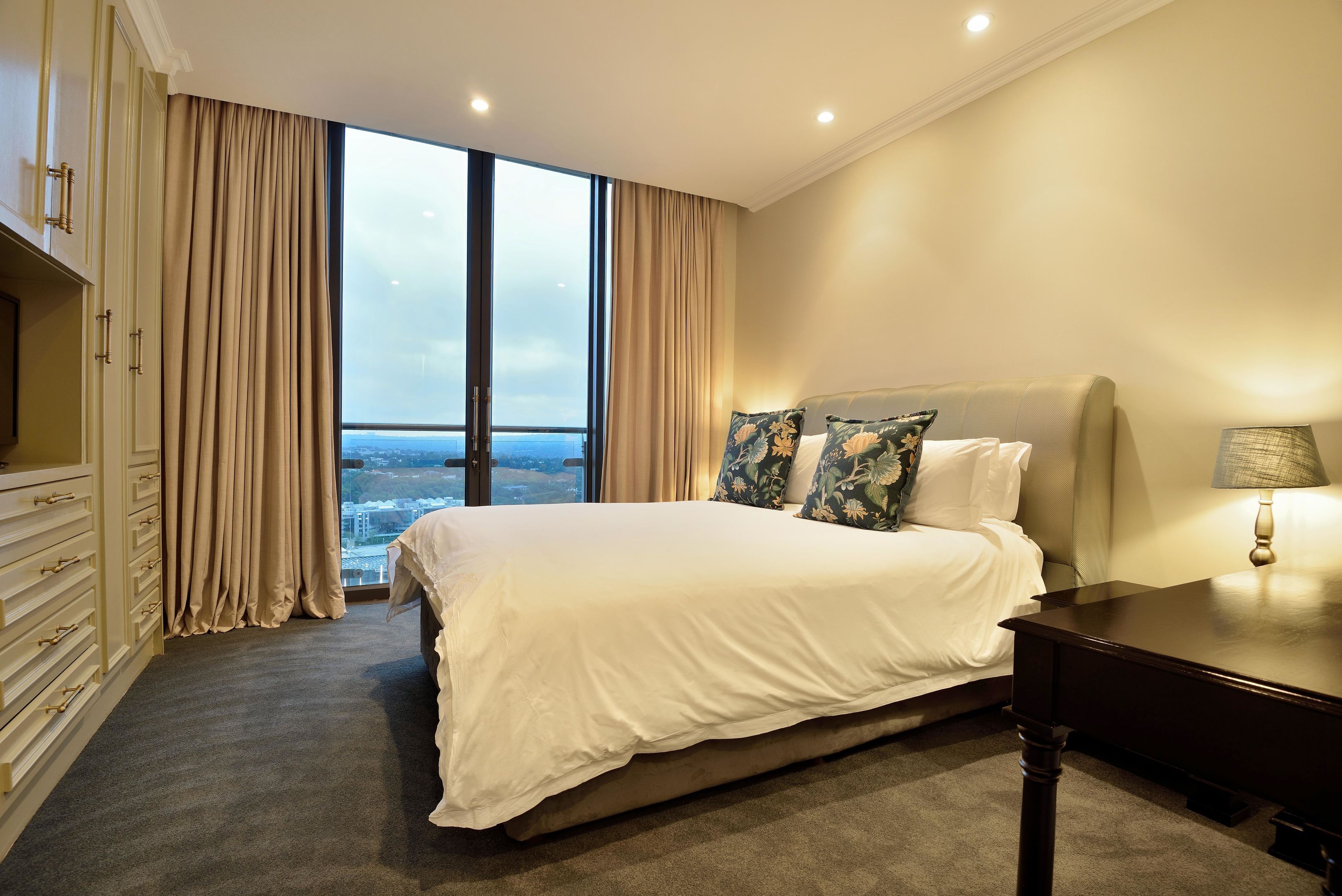 Un dormitorio espacioso con una cama grande, una mesita de noche y vistas a la ciudad a través de ventanales de gran altura.