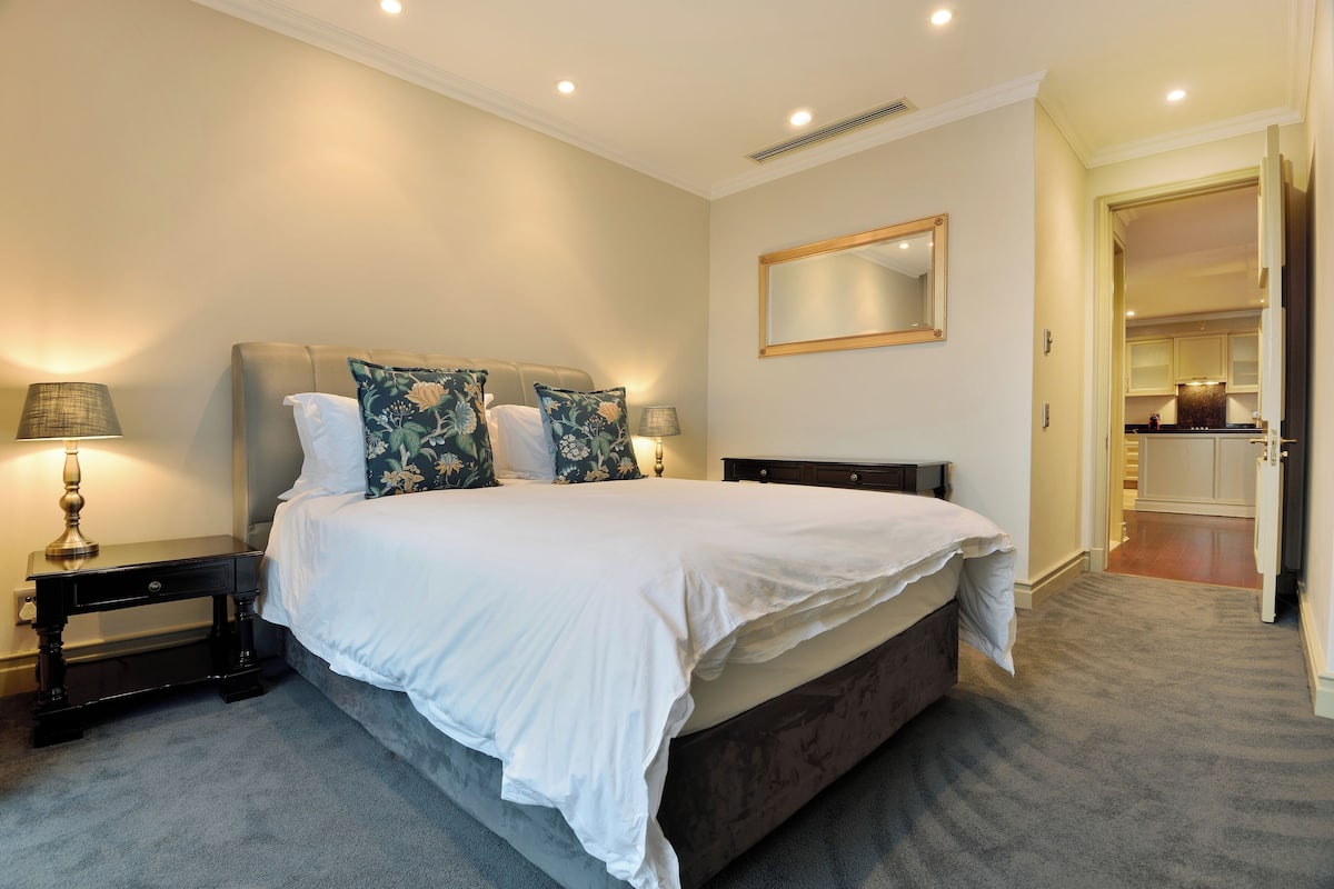 Een slaapkamer met een groot bed, een nachtkastje met een lamp en een spiegel aan de muur.