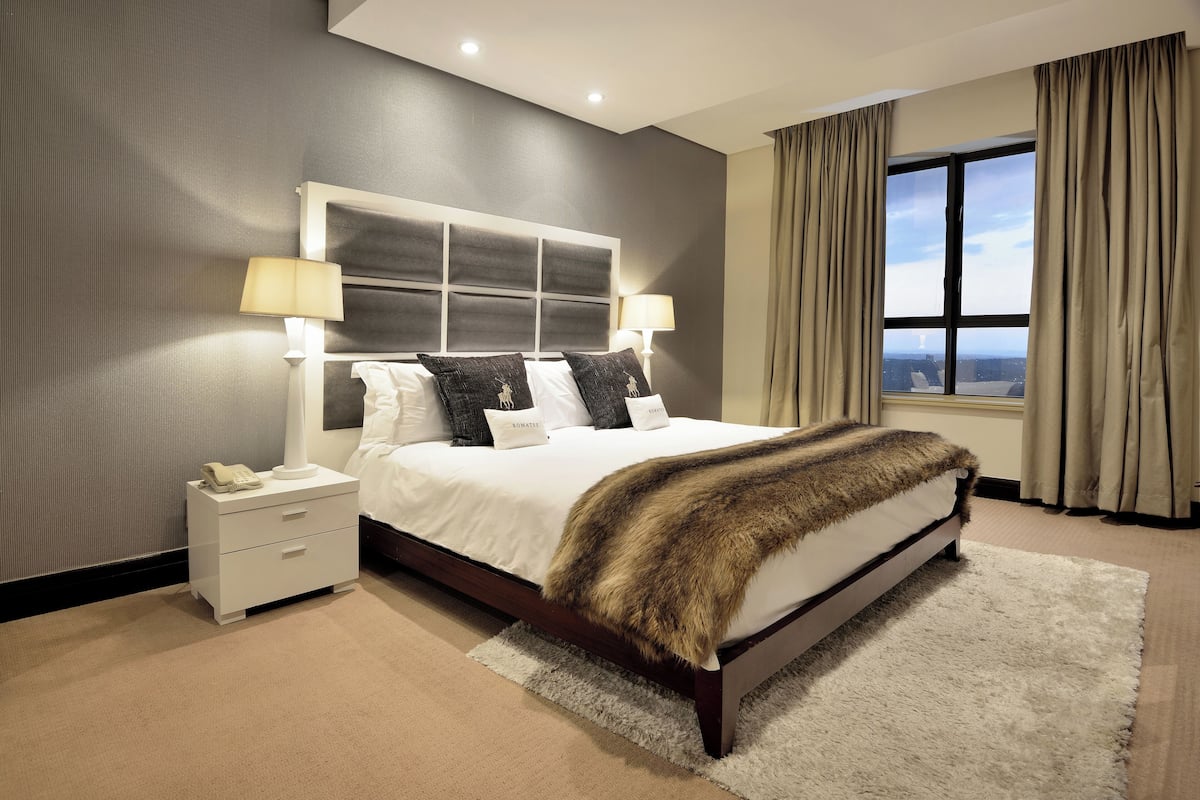 Een moderne slaapkamer met een groot bed, twee bedlampen, een nachtkastje met een telefoon en uitzicht op de stad door een groot raam.