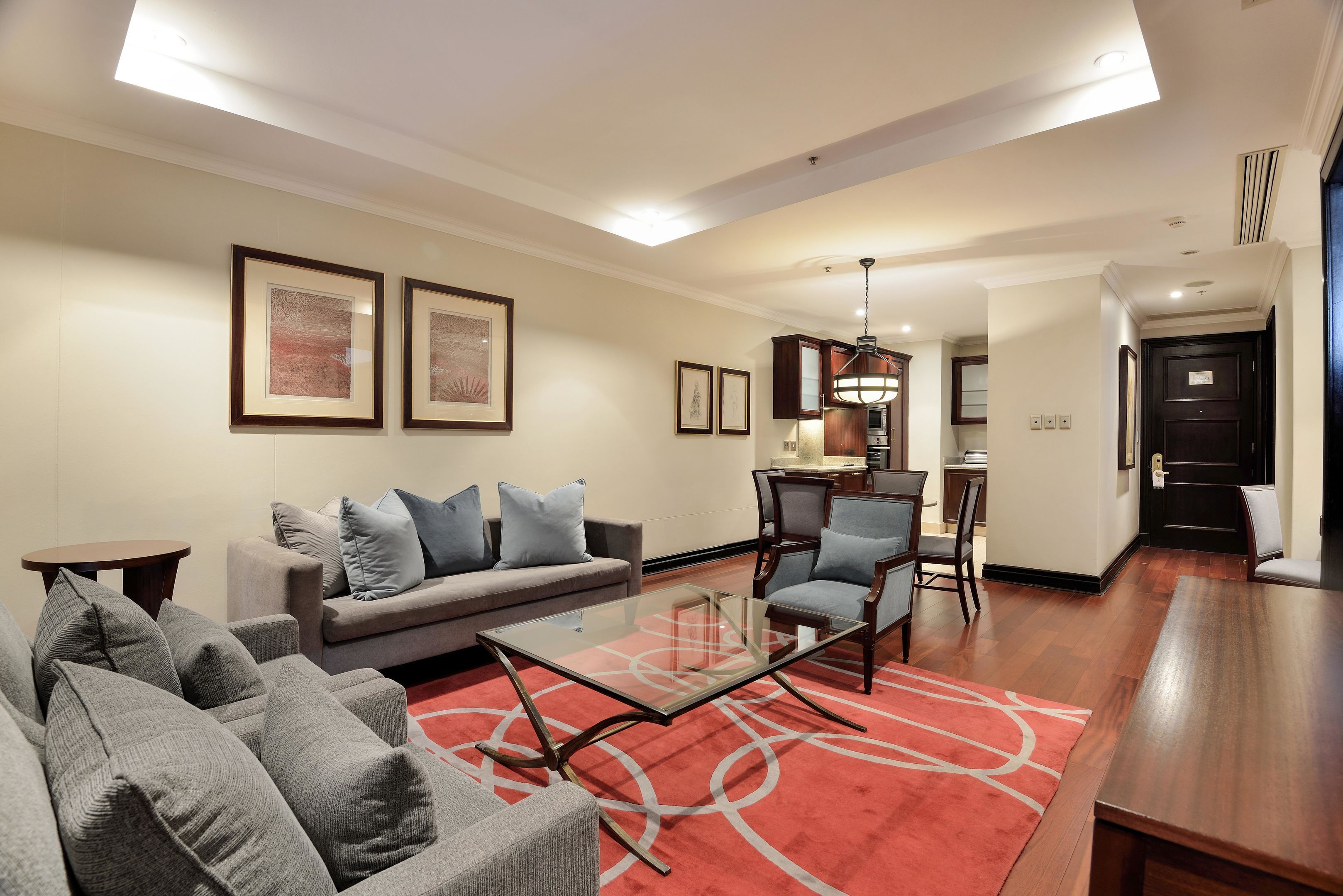 A modern living room with a sofa, armchairs, a glass coffee table, and a red carpet. There are framed pictures on the wall and a chandelier.