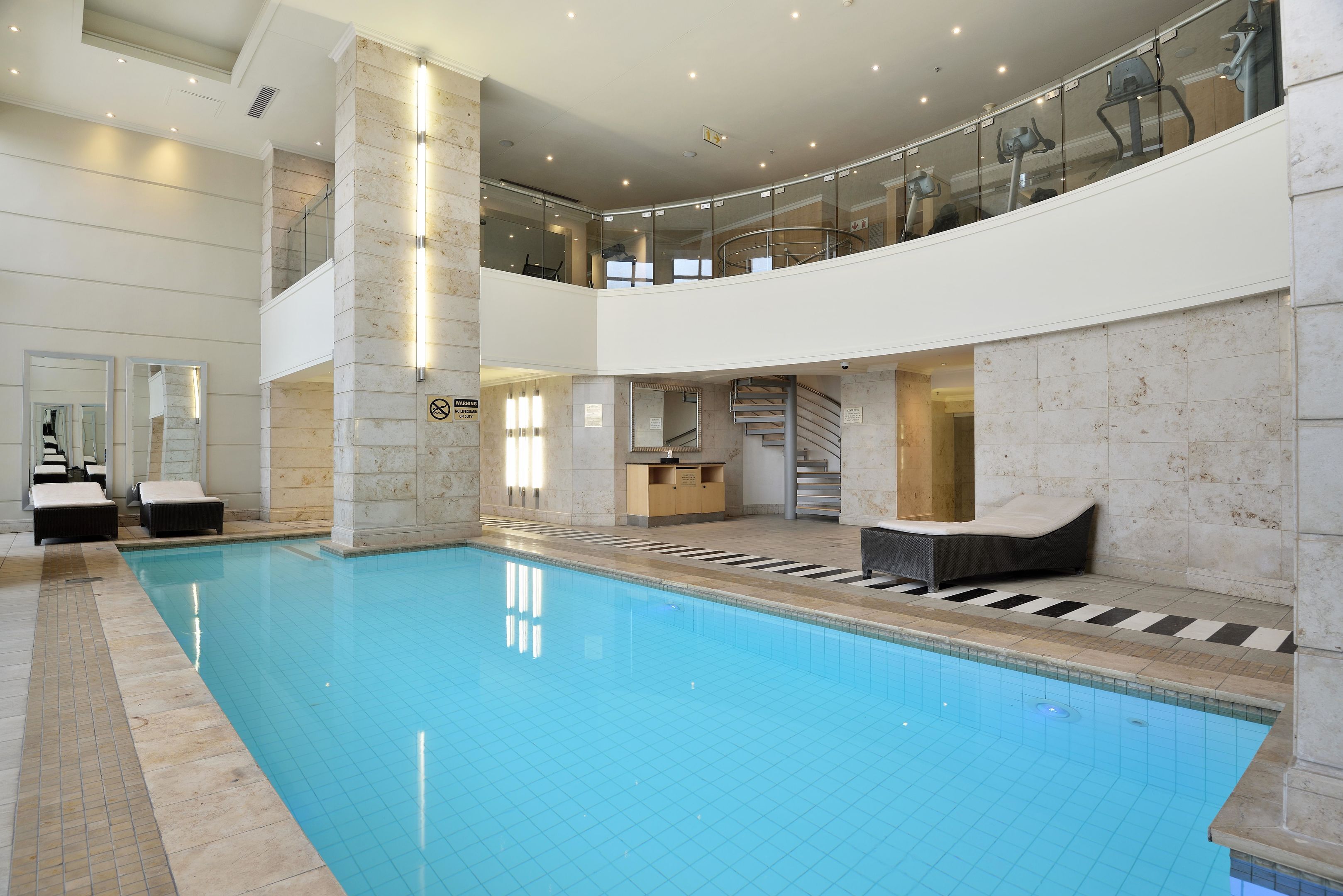 A large indoor swimming pool with a glass balustrade, a staircase, and lounge seating.