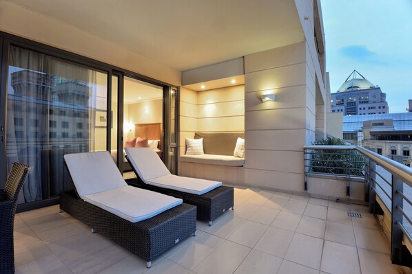 A balcony with two lounge chairs and a cushioned bench, overlooking a cityscape.