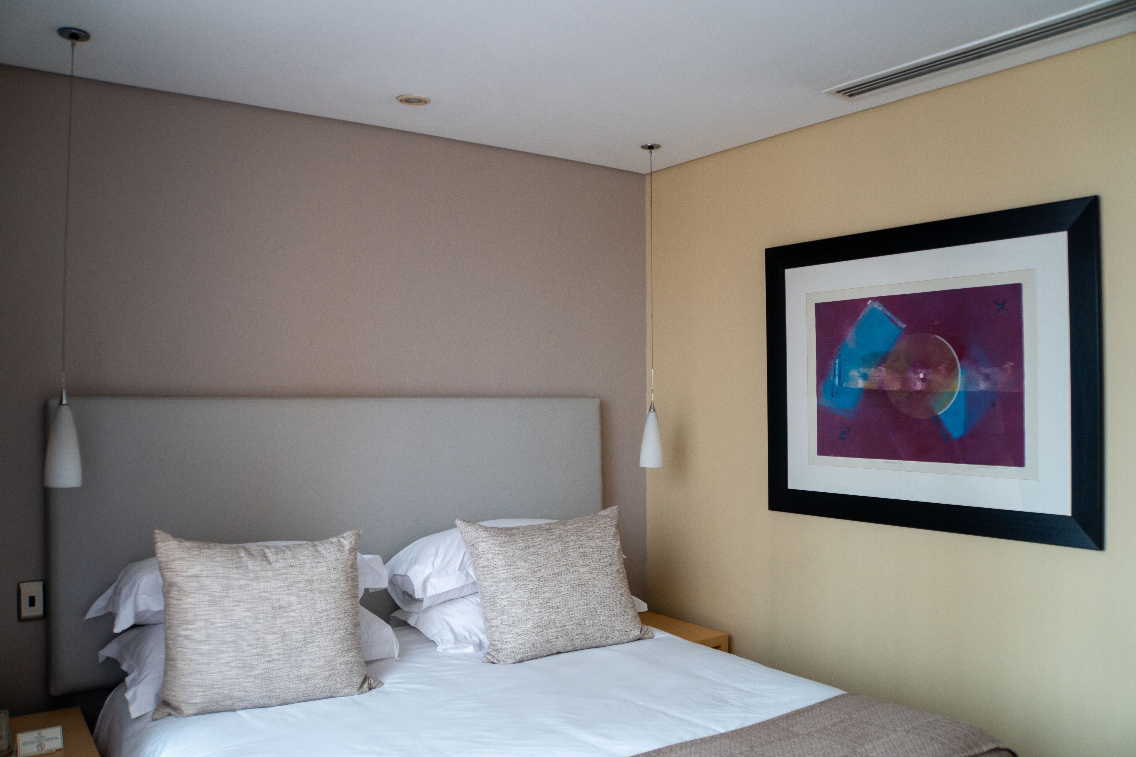 Een keurig opgemaakt bed met witte lakens en twee kussens, een nachtkastje met een lamp, en een lijst met een abstract schilderij aan de muur.