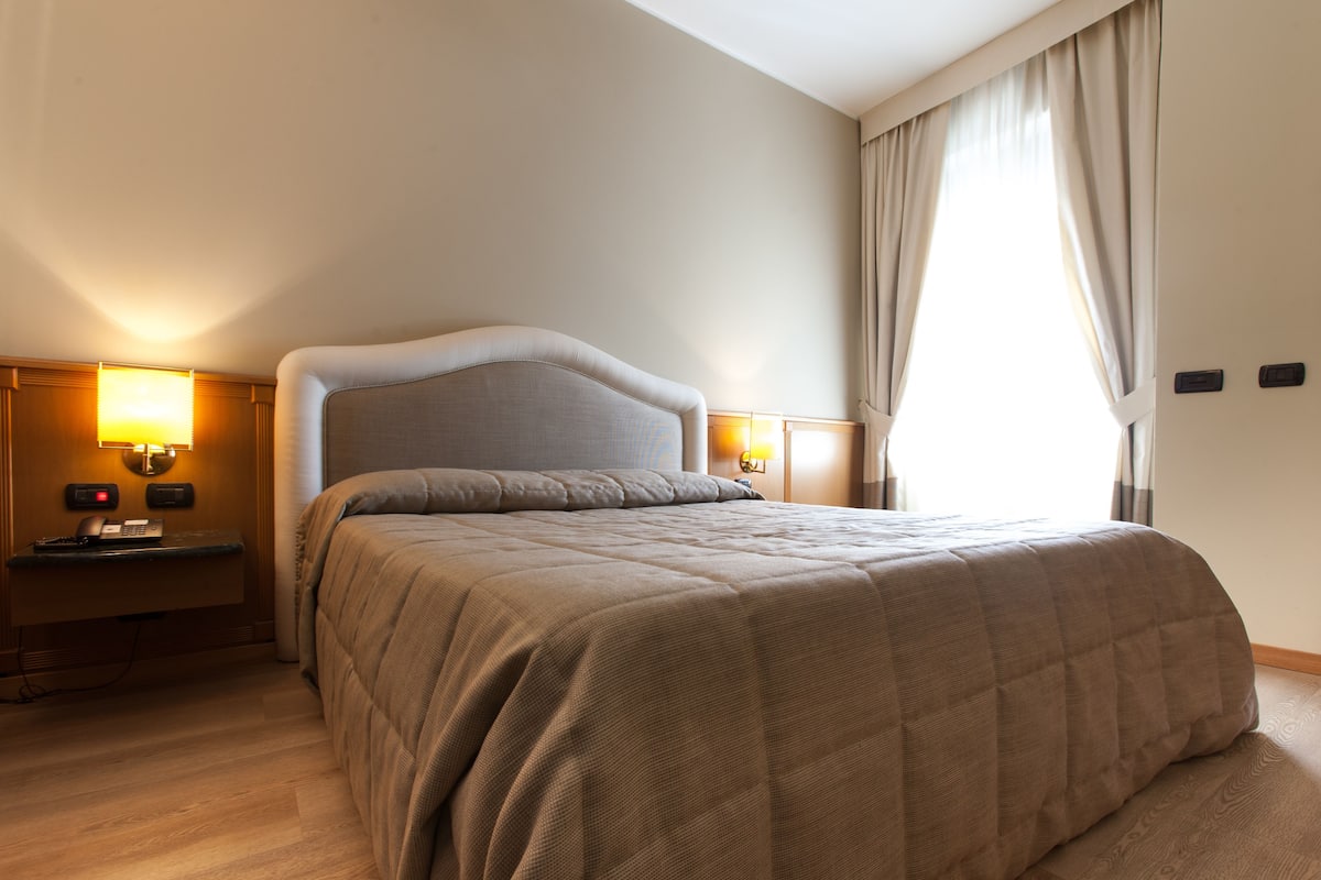 Camera d'albergo con un grande letto, abat-jour, comodino e finestra con tende.