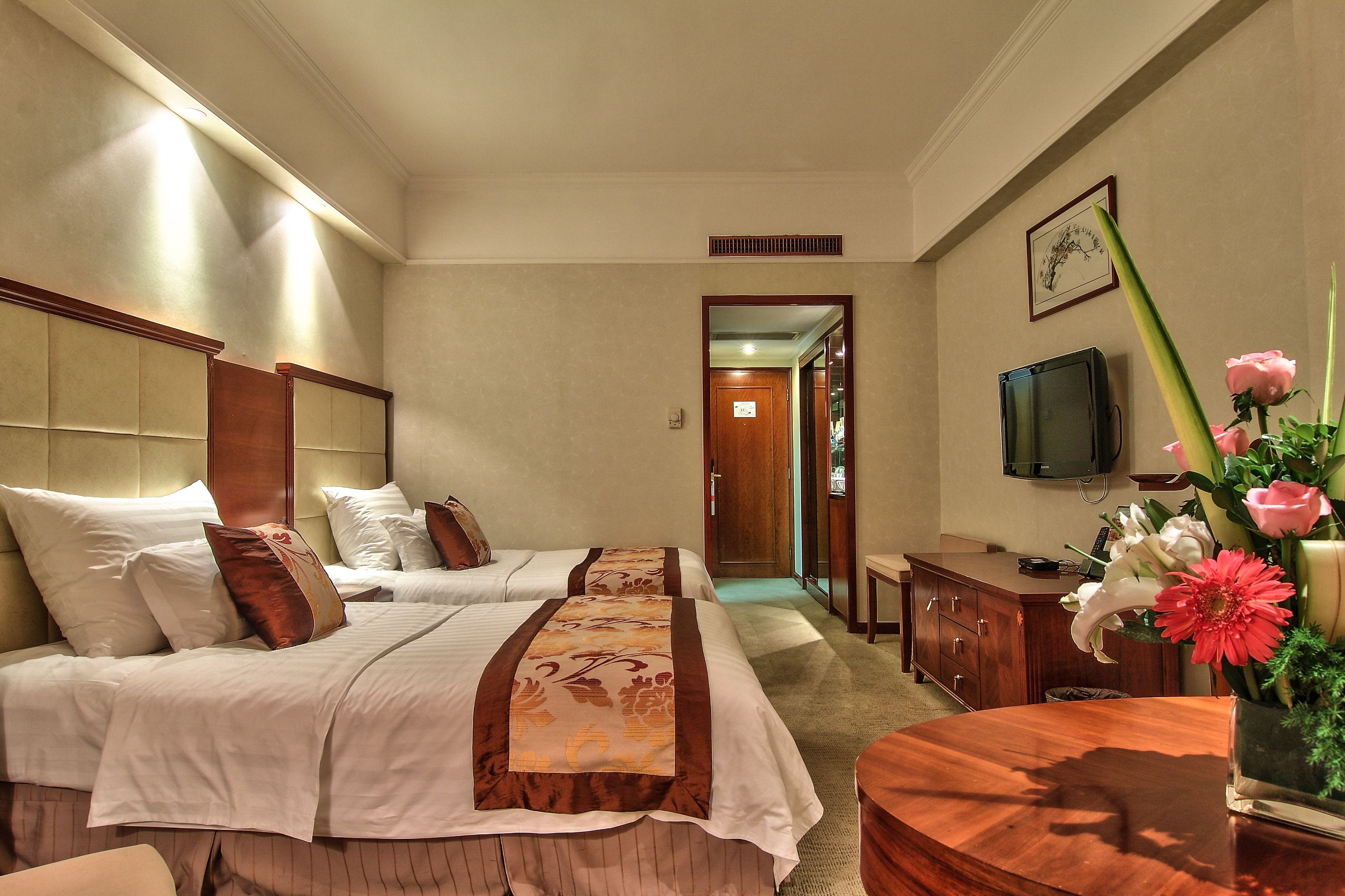 A hotel room with two beds, a wooden desk, a TV, and a vase with flowers.