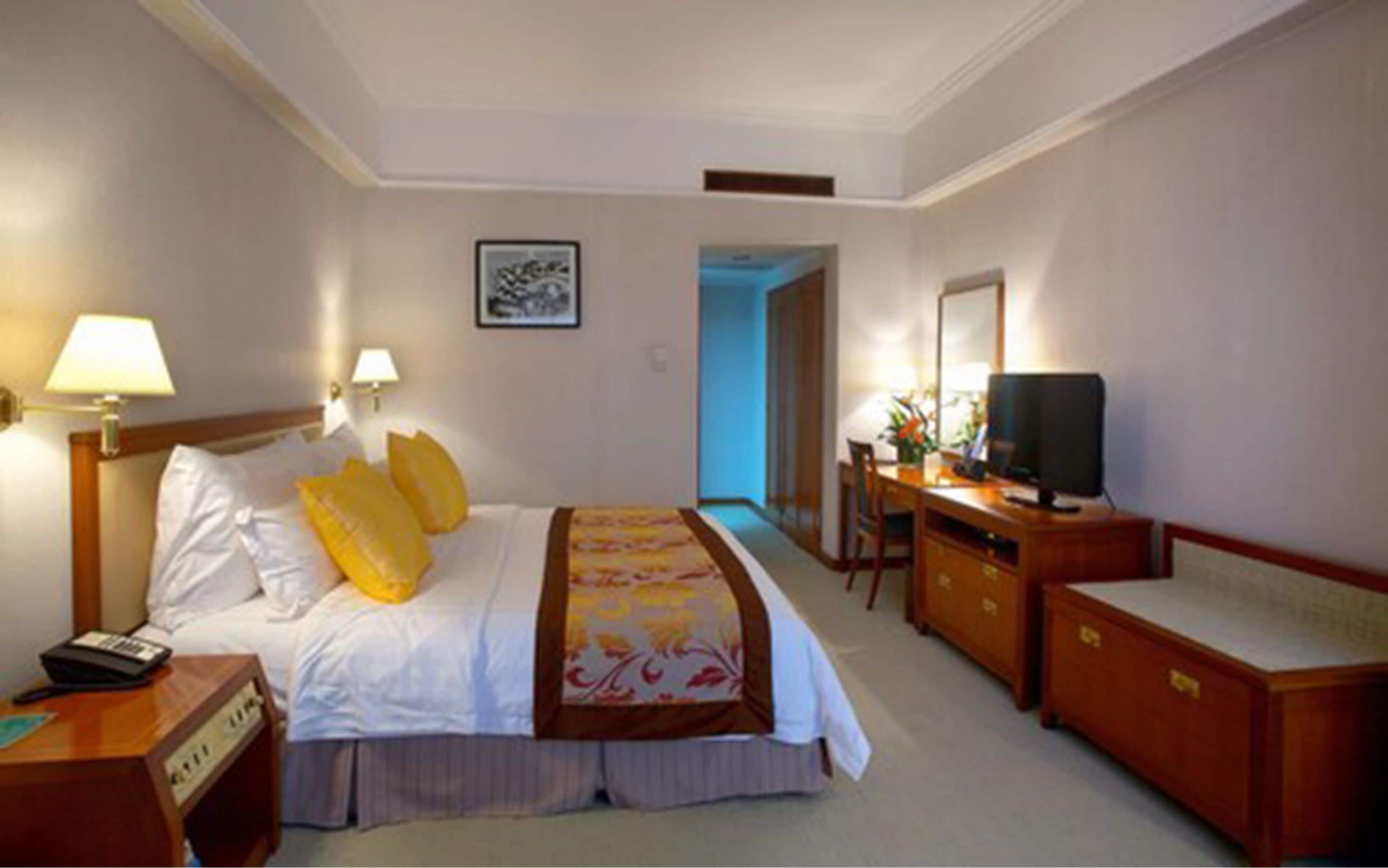 A hotel room with a bed, bedside lamps, a desk with a television, and a dresser.