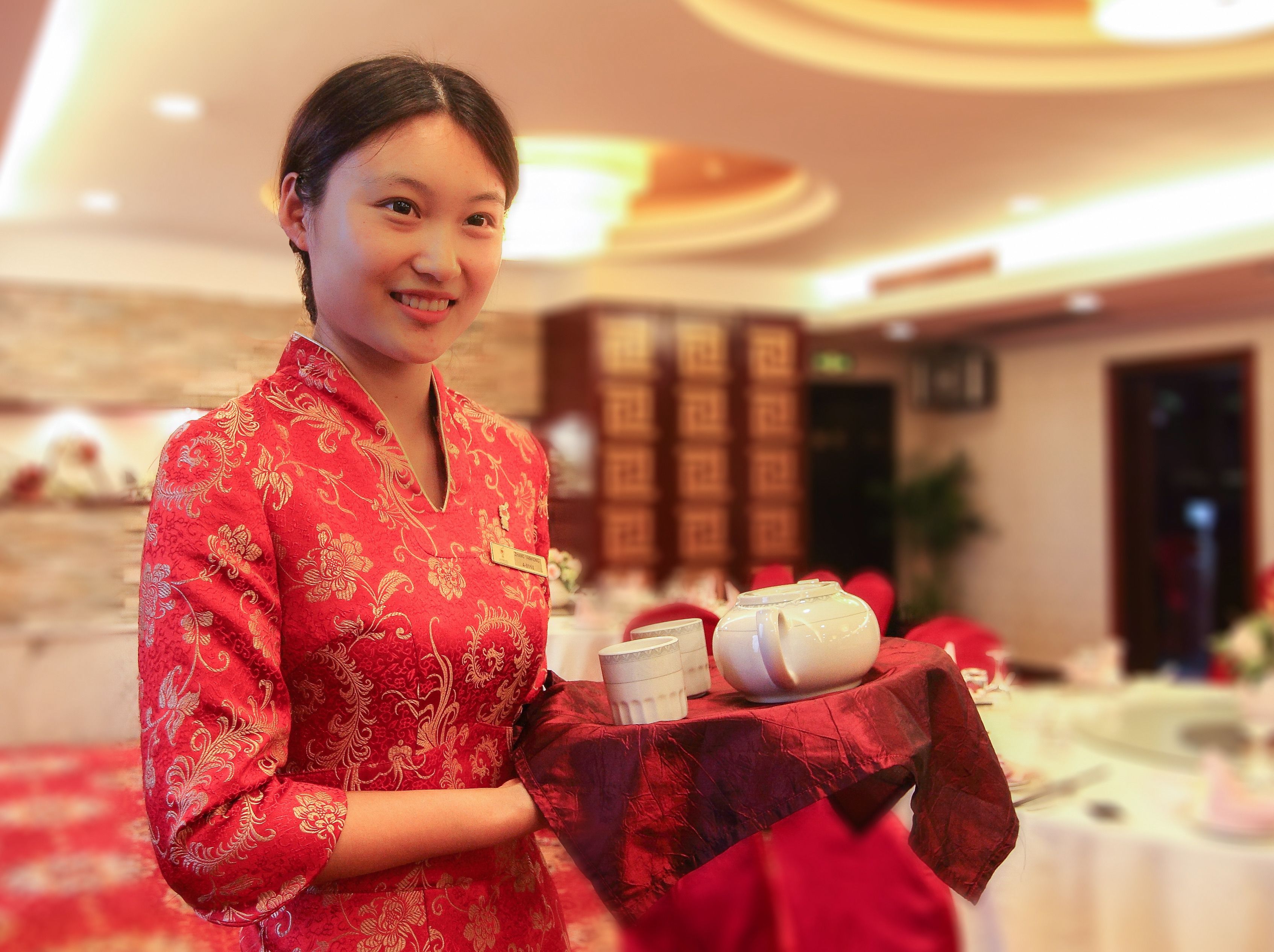 A person in a red traditional outfit holding a tray with teapots and cups, in a room with a red carpet and a table with a white tablecloth.