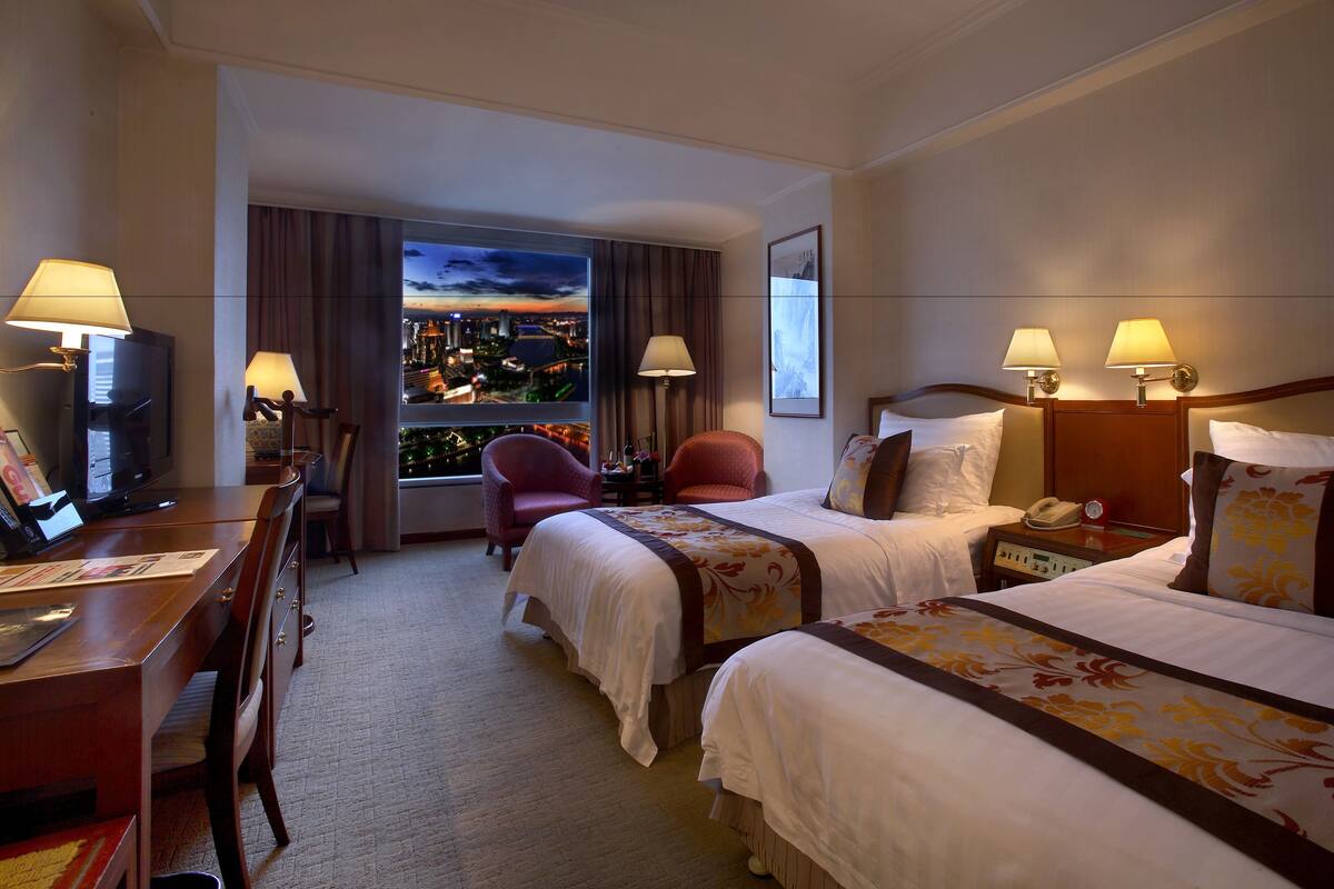 Een hotelkamer met twee bedden, een bureau met een televisie, een stoel en een raam met uitzicht op de stad.