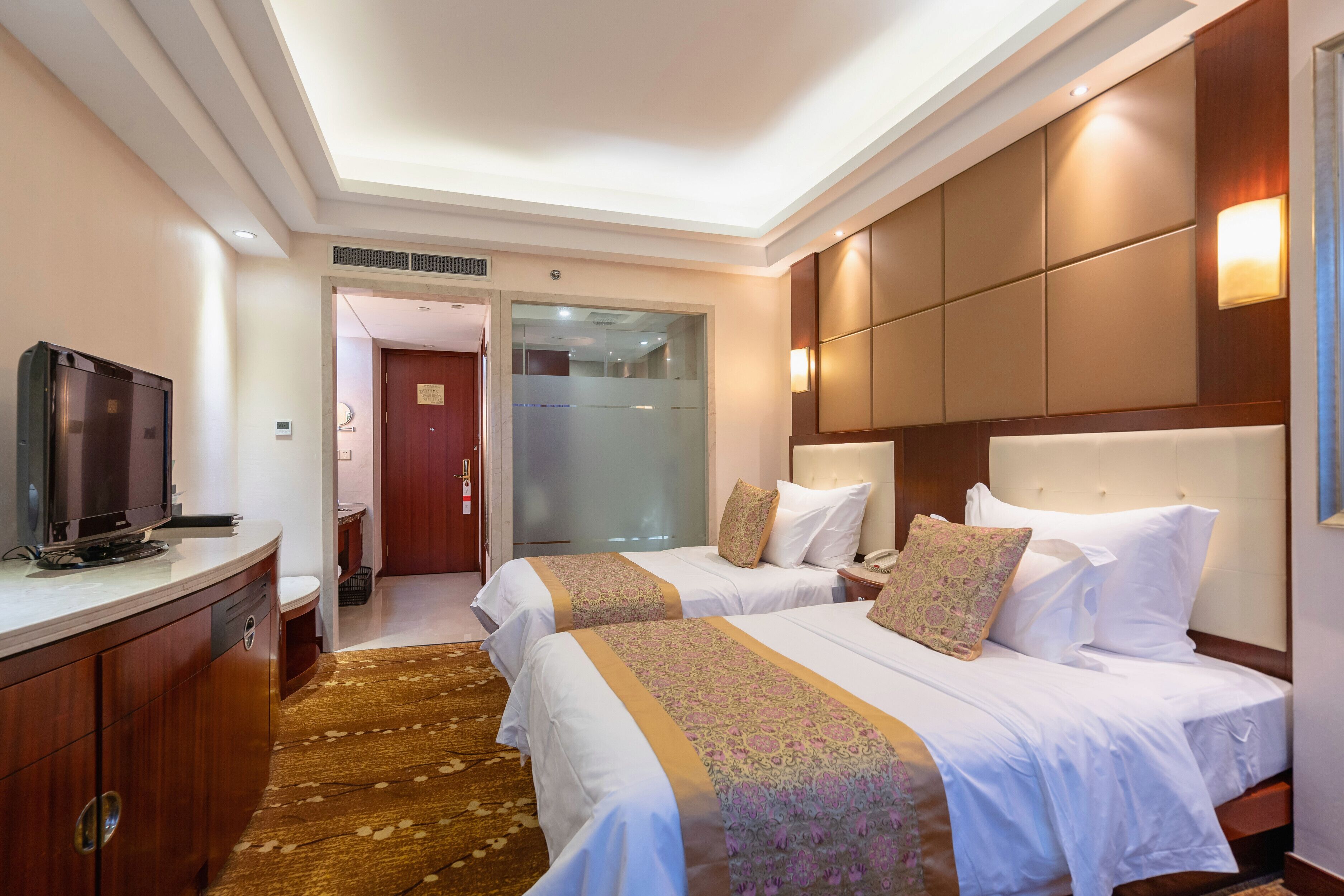Une chambre d’hôtel avec deux lits, un téléviseur posé sur une commode en bois, un sol recouvert de moquette et une porte en verre donnant sur la salle de bain.