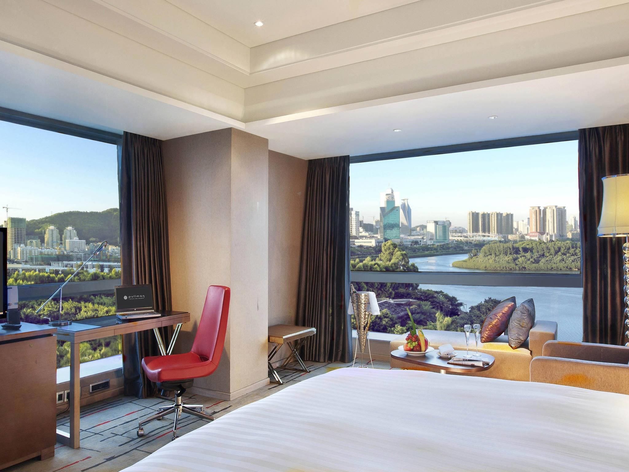 A hotel room with a large bed, a desk with a computer, a red chair, a sofa, and a view of a cityscape and river.