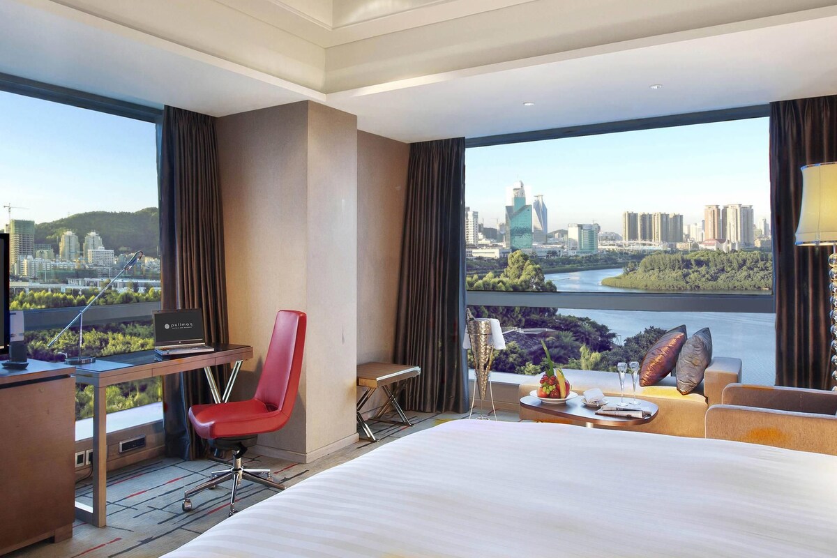 A hotel room with a large bed, a desk with a computer, a red chair, a sofa, and a view of a cityscape and river.