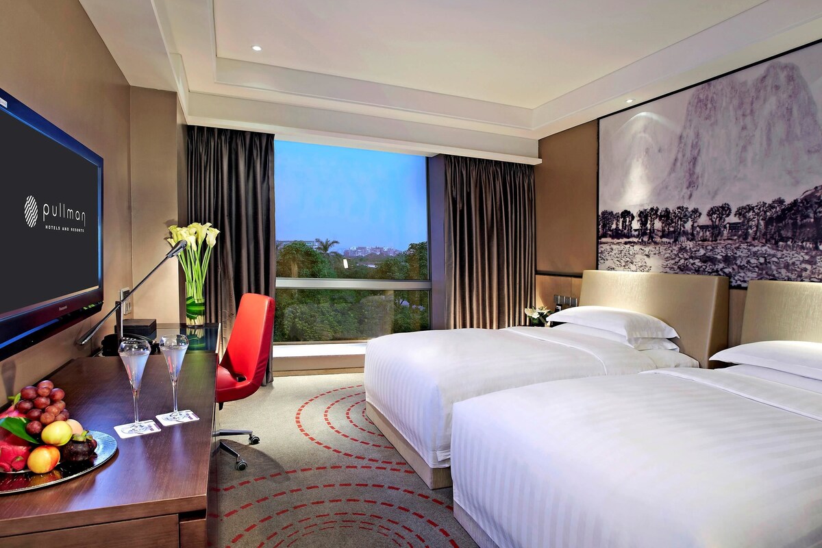 A hotel room with a bed, a desk with a TV, a chair, a window with curtains, and a view of the city.