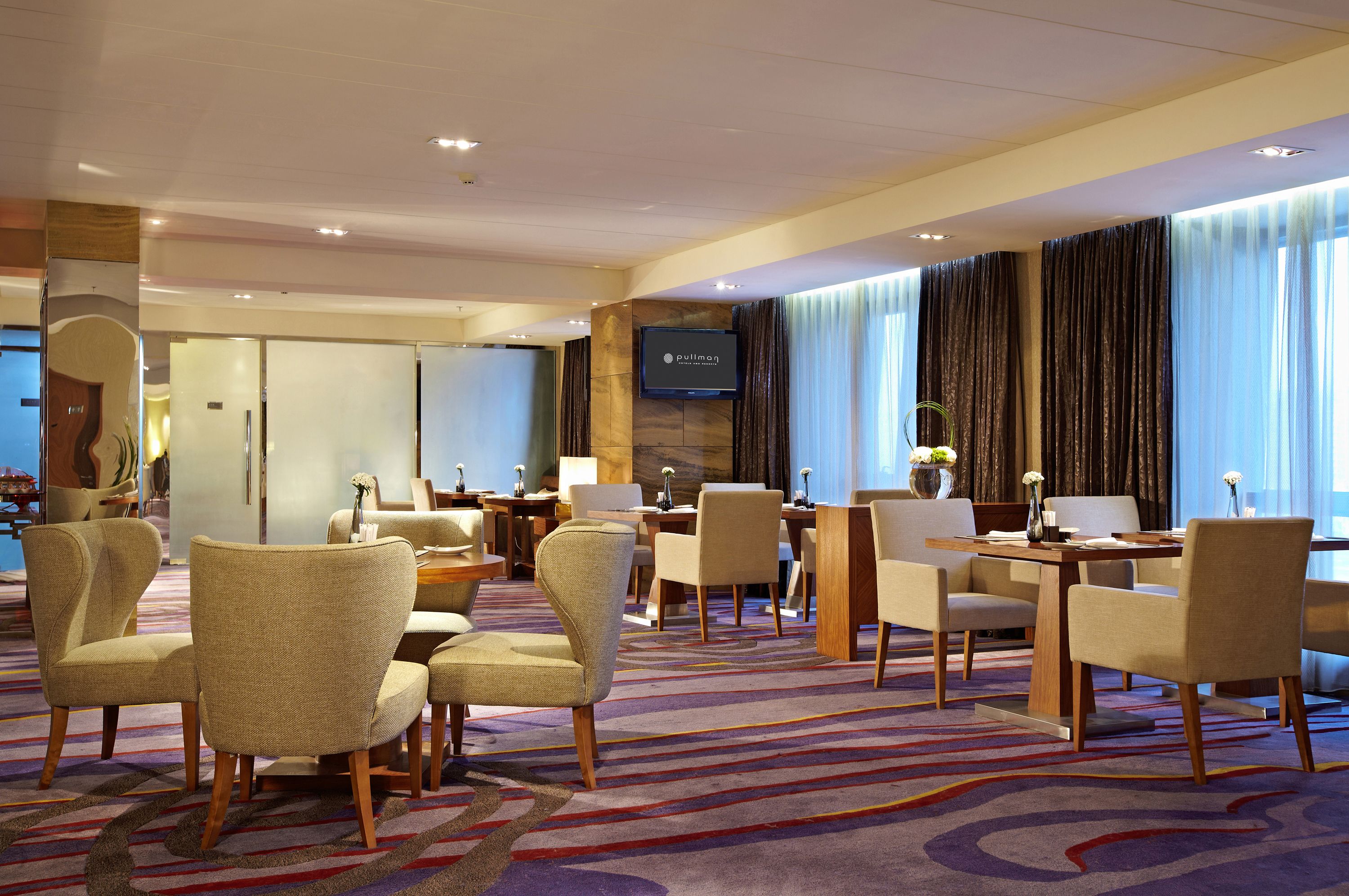 Hall d'albergo con diverse zone salotto, uno schermo televisivo e ampie finestre con tende.
